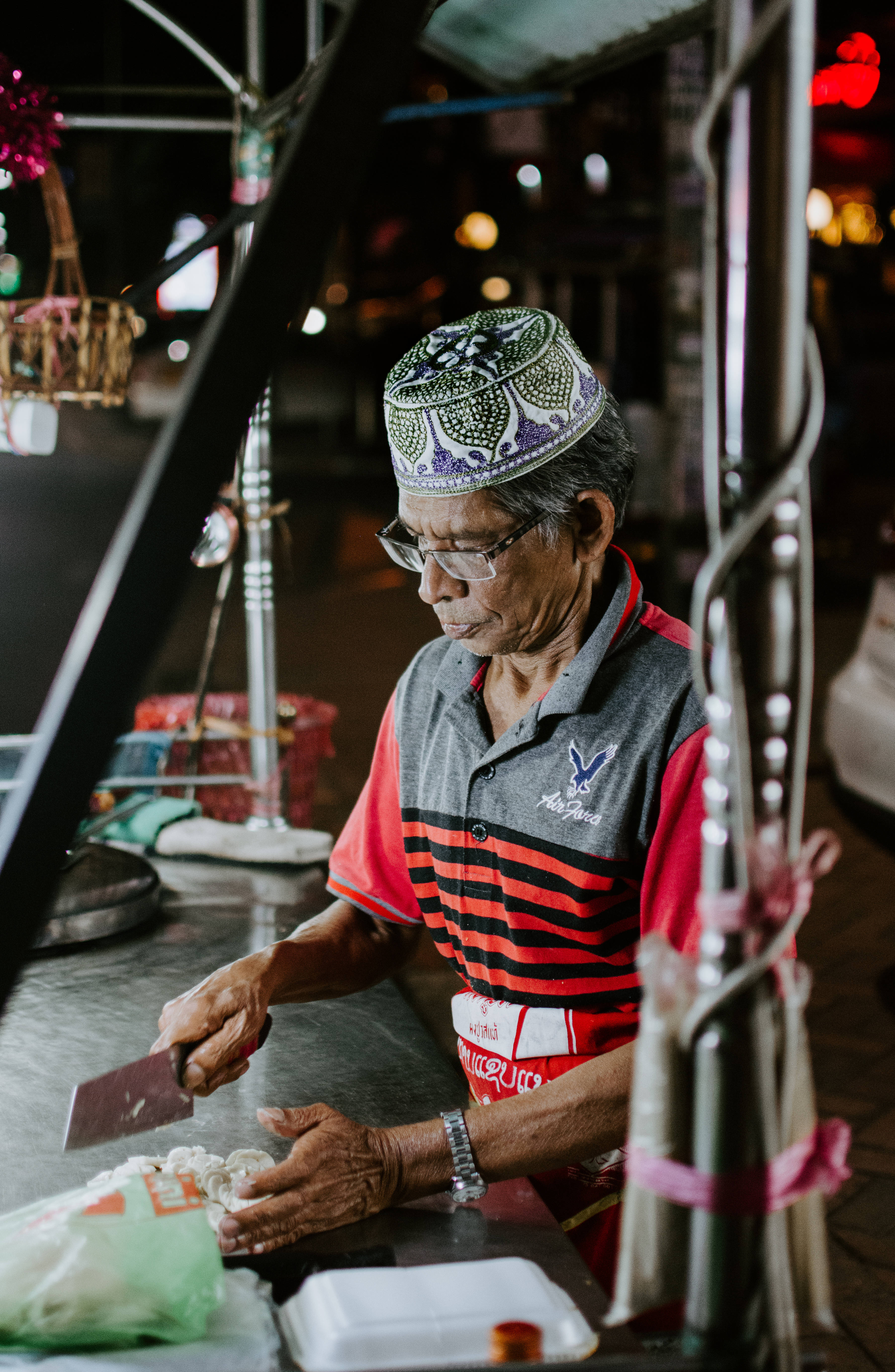 Laos 桌布 壁紙 — 一位戴著圖案帽子和眼鏡的長者，在熱鬧的夜市攤位小心地切食物。