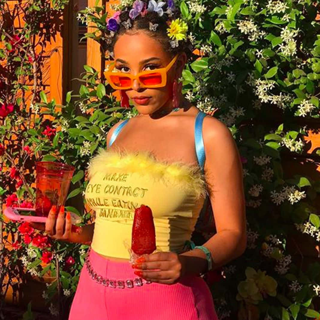 Doja Cat 桌布 壁紙 — 一位戴著大膽橙色太陽眼鏡和黃色羽毛上衣的女性，在茂盛的花卉背景下拿著紅色冰棒和飲料。