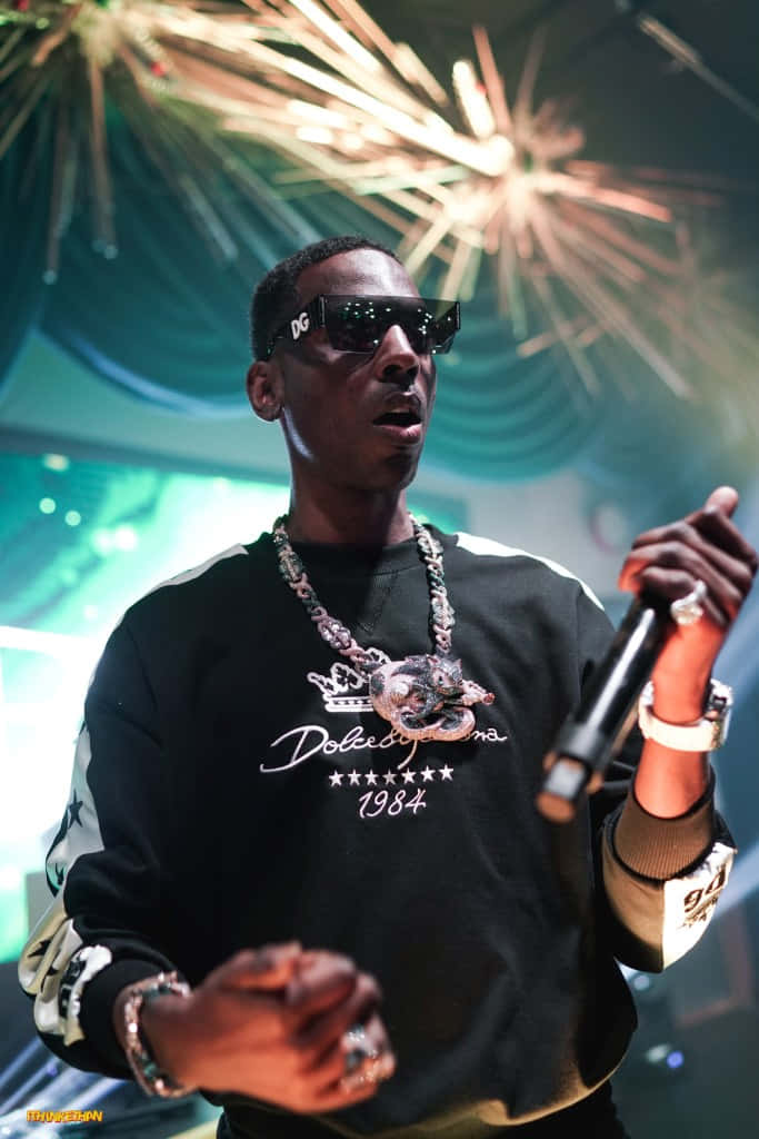 Young Dolph 桌布 壁紙 — 一位戴著太陽眼鏡、身穿 Dolce & Gabbana 連帽衫的饒舌歌手，在耀眼的舞台煙火下表演。