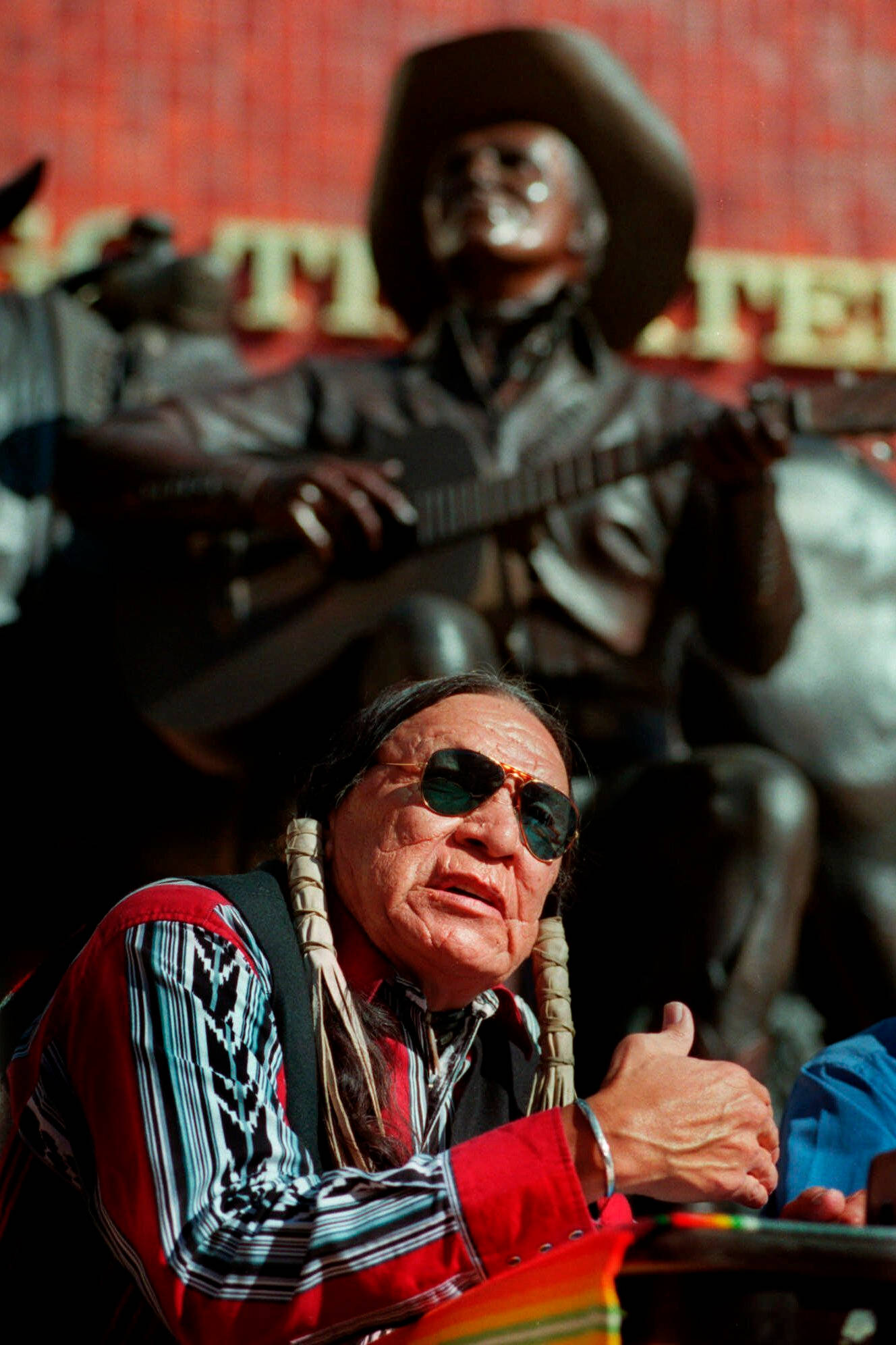Native American 桌布 壁紙 — 一位戴著太陽眼鏡、身穿傳統服飾的長者美洲原住民男子，在一尊青銅牛仔雕像前豎起大拇指。
