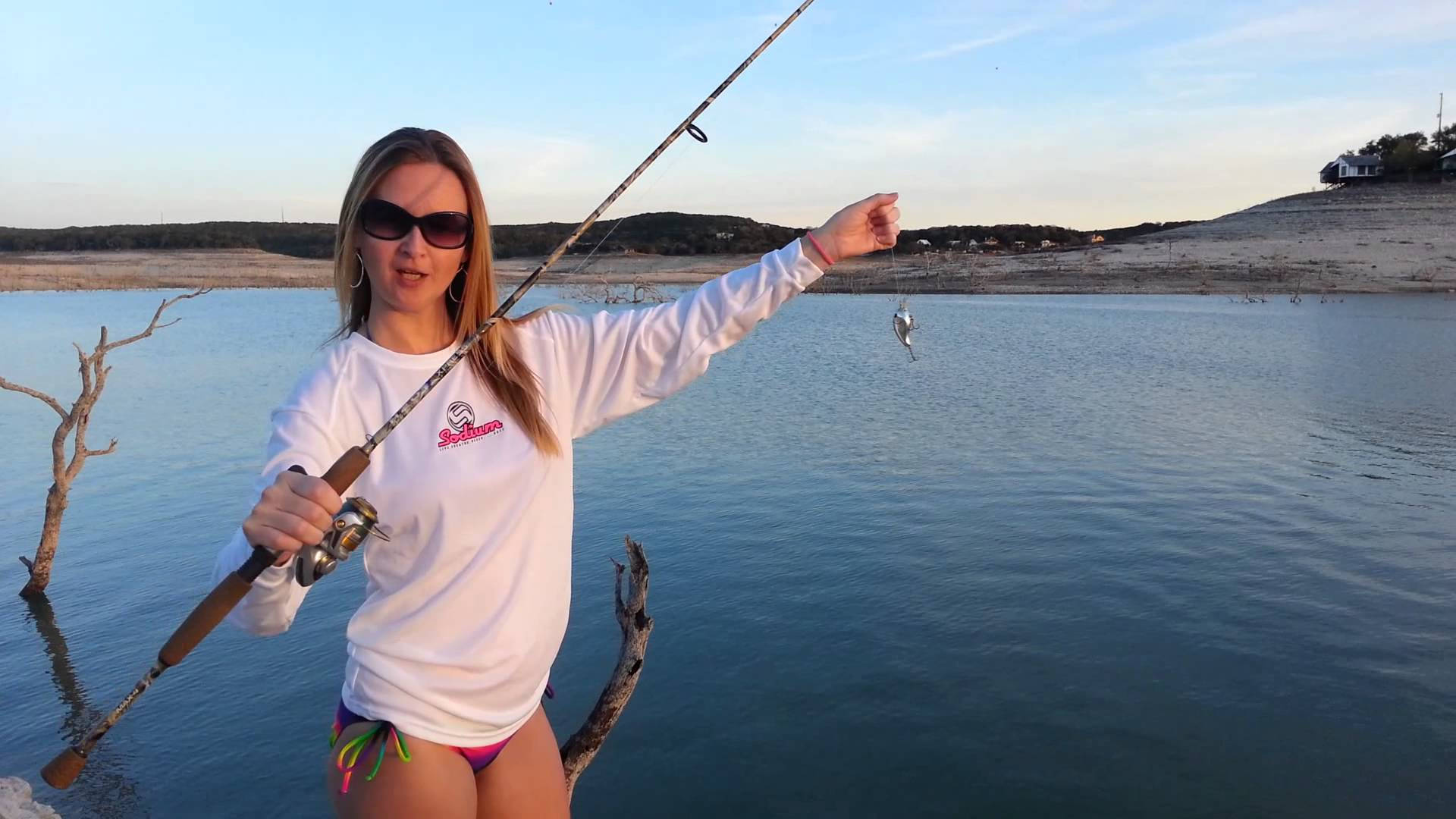 釣魚 桌布 壁紙 — 一位戴著太陽眼鏡、身穿白襯衫的女性，在日落時分的平靜水邊自豪地展示釣竿上的一條小魚。