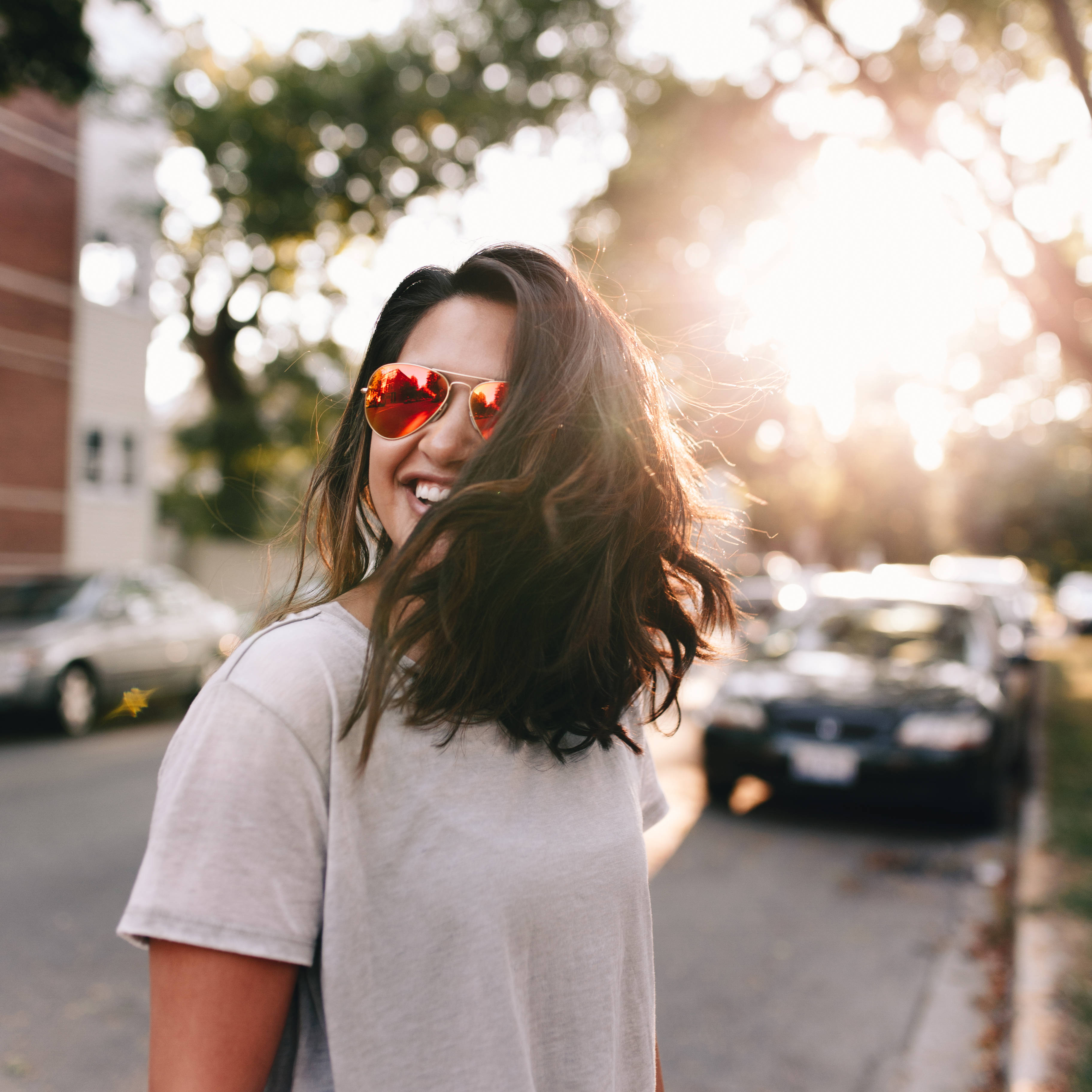 Smile 桌布 壁紙 — 一位戴著太陽眼鏡、頭髮被風吹拂的微笑女士站在陽光明媚的街道上，身後有汽車和樹木，呈現柔焦效果。