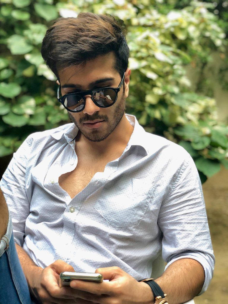 Feroz Khan 桌布 壁紙 — 一位戴著太陽眼鏡和淺藍色襯衫的時尚男士在茂密的綠色樹葉中專注地看著手機。