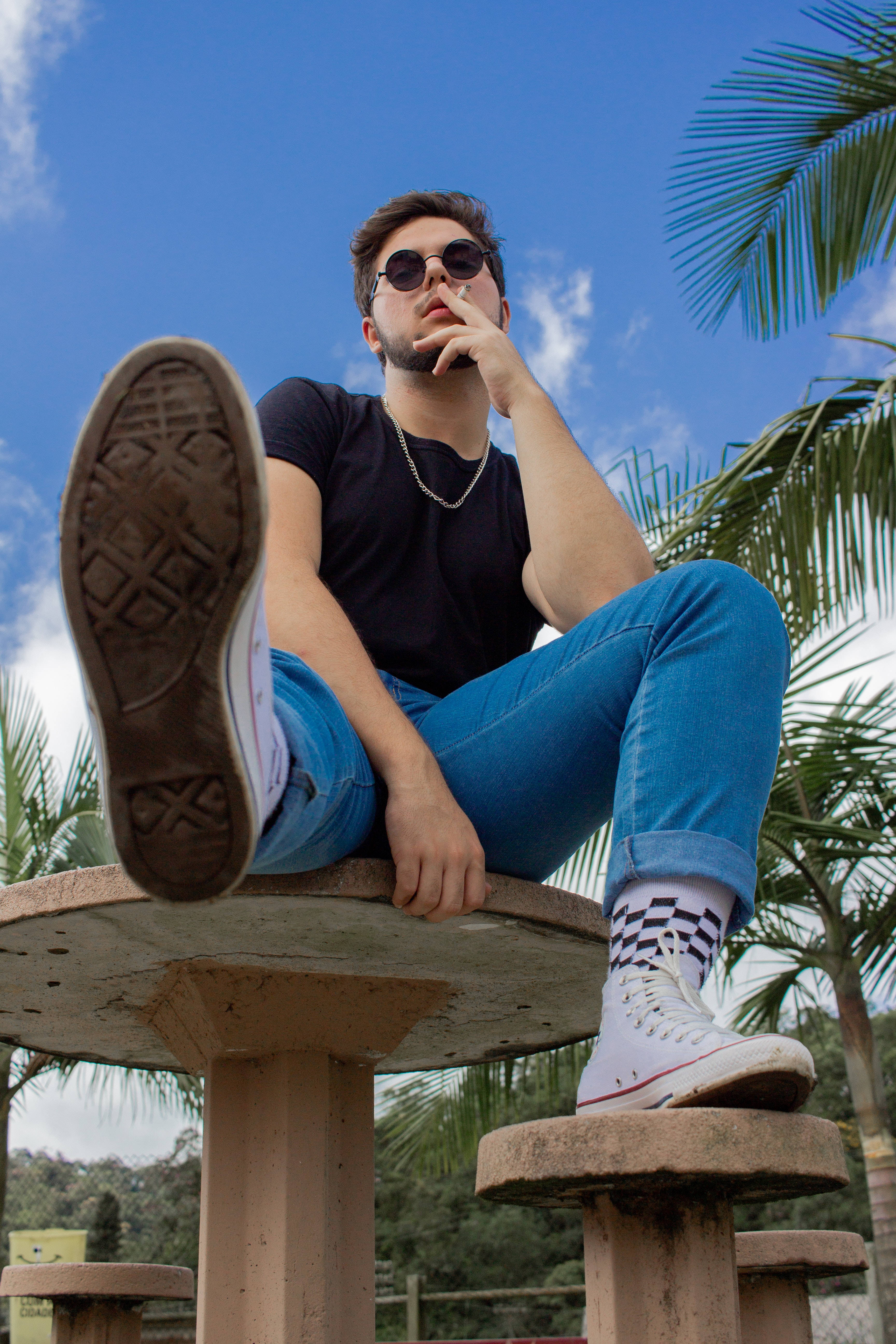 male model 桌布 壁紙 — 一位戴著太陽眼鏡和牛仔褲的年輕男子，躺在一座木結構上，翹起一隻腳，周圍環繞著棕櫚樹，頭頂是明亮的藍天。