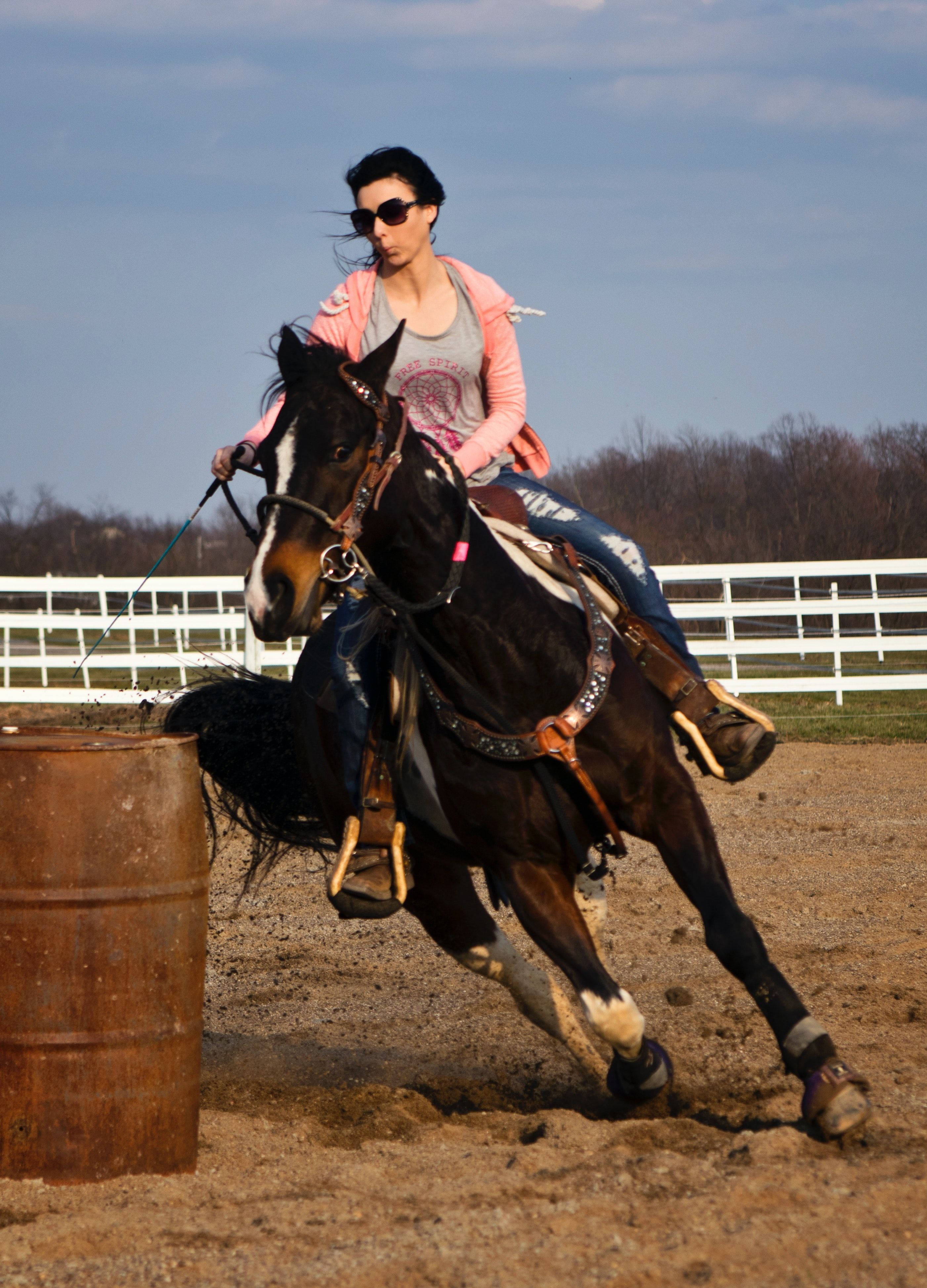 Barrel Racing 桌布 壁紙 — 一位戴著太陽眼鏡和粉紅色外套的騎手，在晴朗的天空下，引導一匹深色馬在圍欄競技場中繞著一個生鏽的桶子急轉彎。