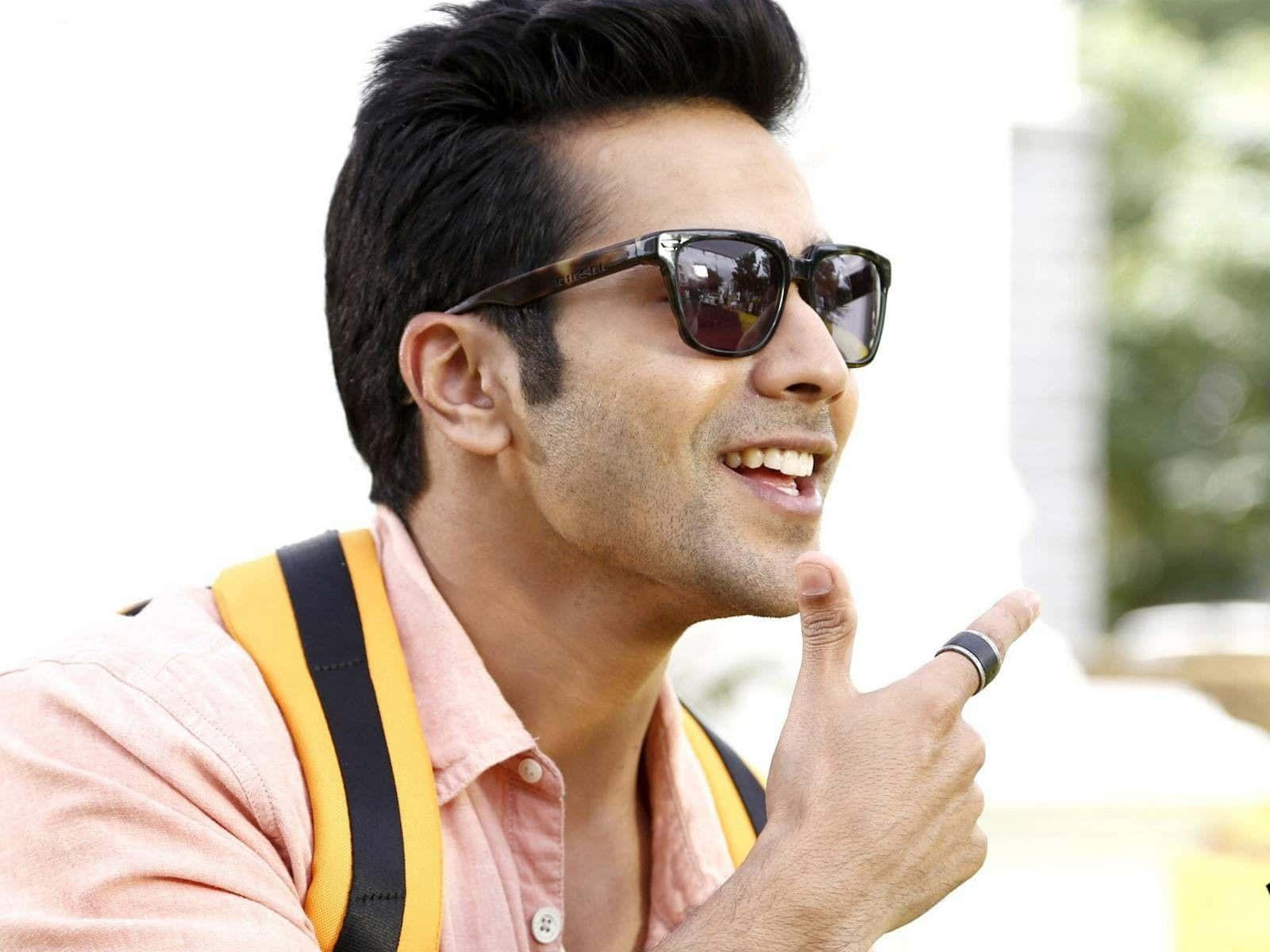 Varun Dhawan 桌布 壁紙 — 一位戴著太陽眼鏡和粉紅色襯衫的微笑男子，背著黃色背包，愉快地指著。