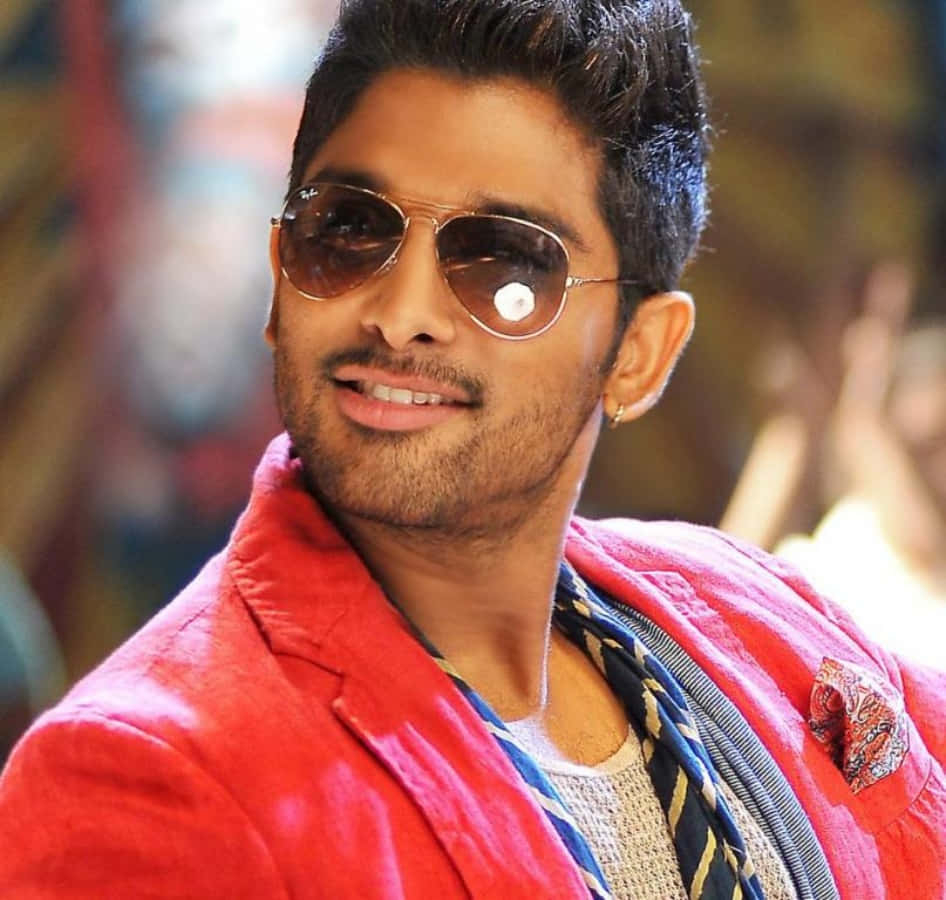 Allu Arjun 桌布 壁紙 — 一位戴著太陽眼鏡和紅色外套的時尚男士，在模糊多彩的背景前自信地微笑。