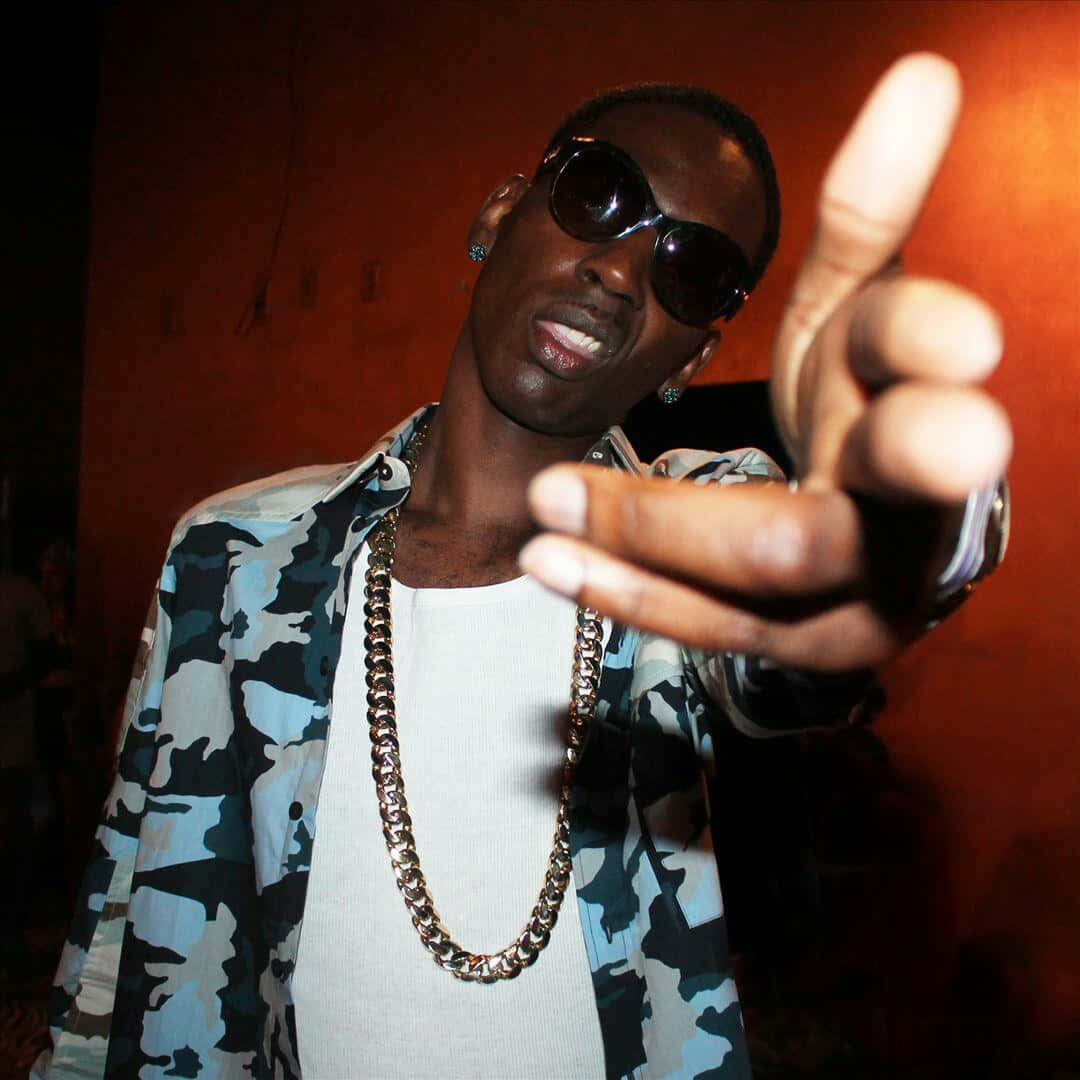 Young Dolph 桌布 壁紙 — 一位戴著太陽眼鏡和迷彩外套的時尚男士，在溫暖的橙色牆壁前自信地指向相機。