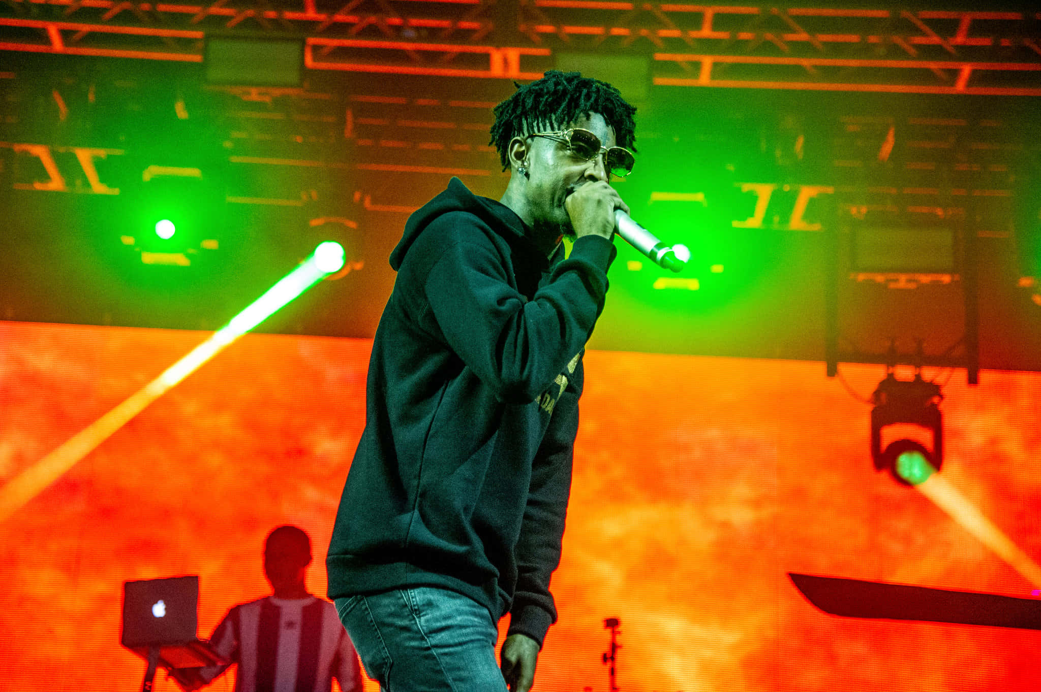 21 Savage 桌布 壁紙 — 一位戴著太陽眼鏡和連帽衫的饒舌歌手在舞台上表演，背景是鮮豔的綠色和橙色燈光，還有一位筆記型電腦技術人員。