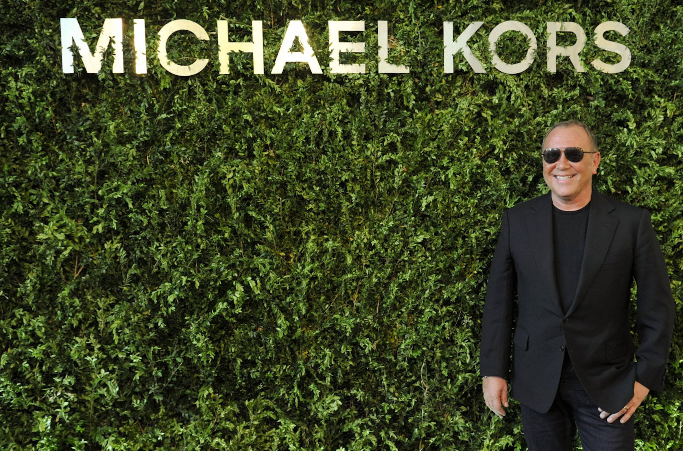 michael 桌布 壁紙 — 一位戴著太陽眼鏡和黑色西裝的微笑男子自信地站在茂密綠色籬笆牆前，牆上有「MICHAEL KORS」標誌。