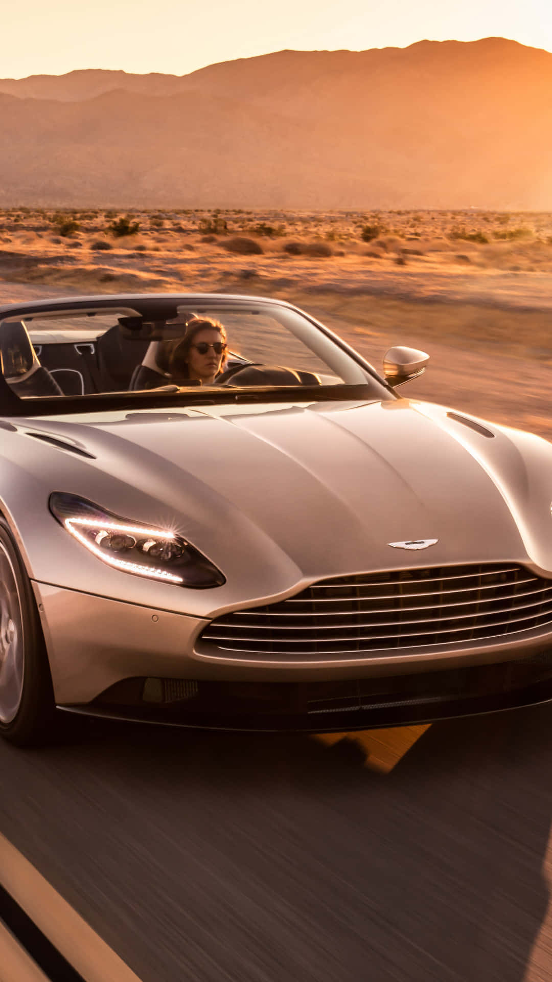 Aston Martin Db11 桌布 壁紙 — 一位戴著太陽眼鏡的女司機駕駛銀色Aston Martin DB11敞篷跑車在日落時分疾馳穿過沙漠。