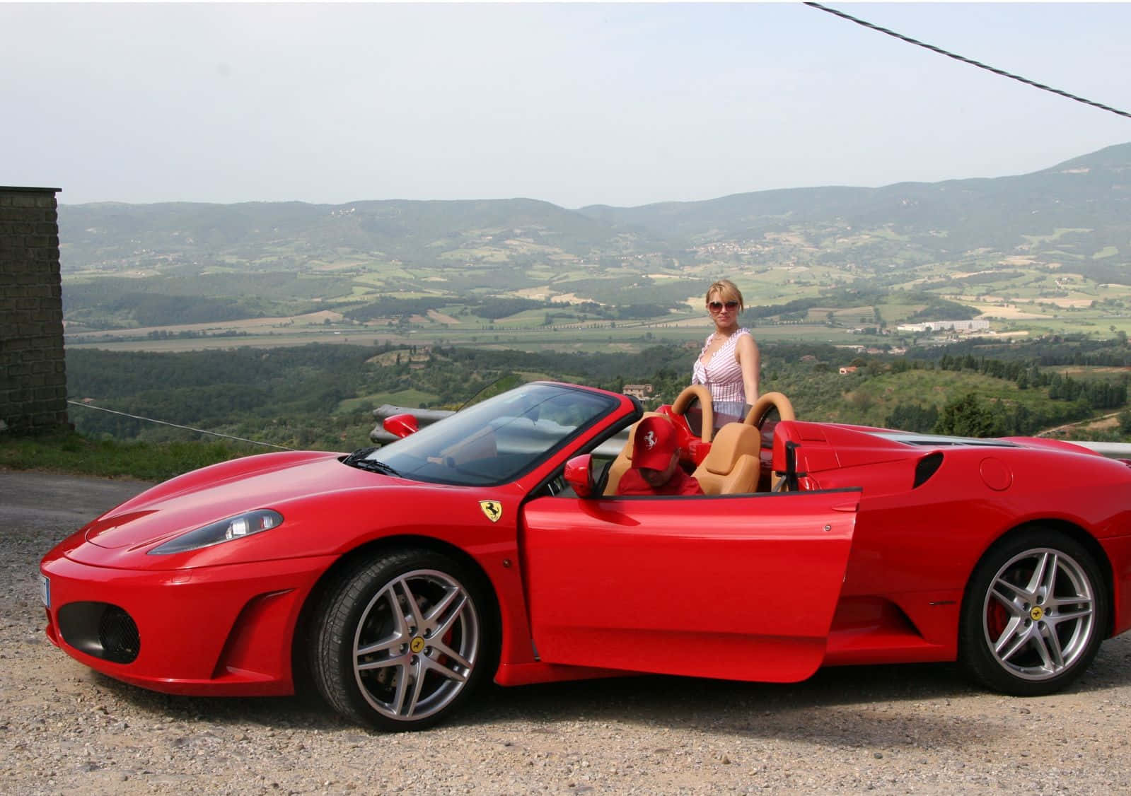 Ferrari F430 桌布 壁紙 — 一位戴著太陽眼鏡的女性站在一輛鮮紅色法拉利敞篷車旁，車停在碎石觀景點上，眼前是起伏的綠色山丘和遠處村莊，天空朦朧。