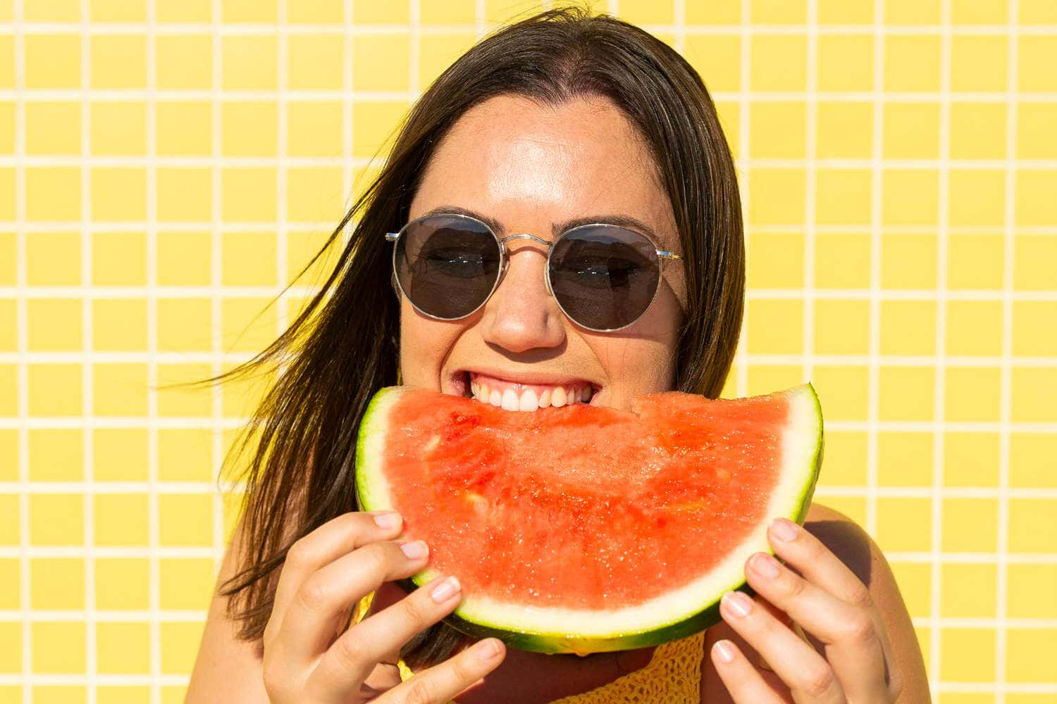 Watermelon 桌布 壁紙 — 一位戴著太陽眼鏡的微笑女士，在明亮的黃色瓷磚牆前愉快地咬著多汁的西瓜片。