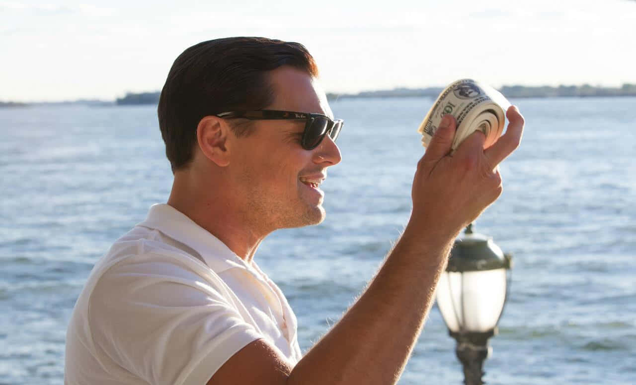 Wolf Of Wall Street 桌布 壁紙 — 一位戴著太陽眼鏡的微笑男子，在明亮的日光下，於海濱燈柱旁拿著一疊捲起的現金。