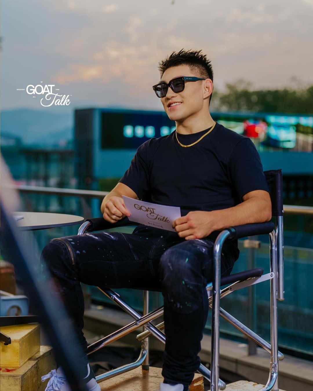 Handsome Boy 桌布 壁紙 — 一位戴著太陽眼鏡的微笑男子自信地坐在導演椅上，手持一張「GOAT Talk」卡片，背景是暮色中的城市景觀。
