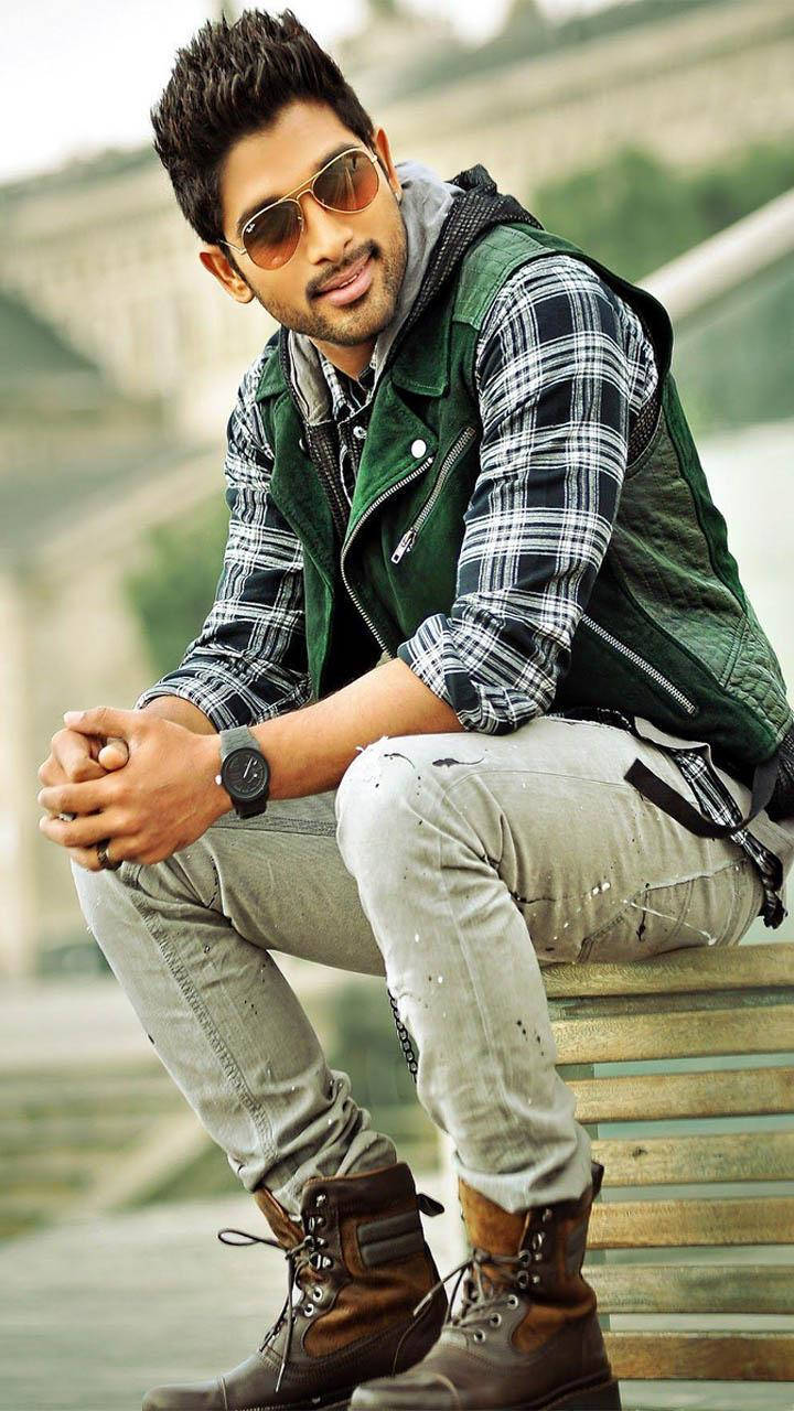 Allu Arjun 桌布 壁紙 — 一位戴著太陽眼鏡的時尚男士自信地坐在木長椅上，身穿格紋襯衫、綠色背心和堅固的靴子。