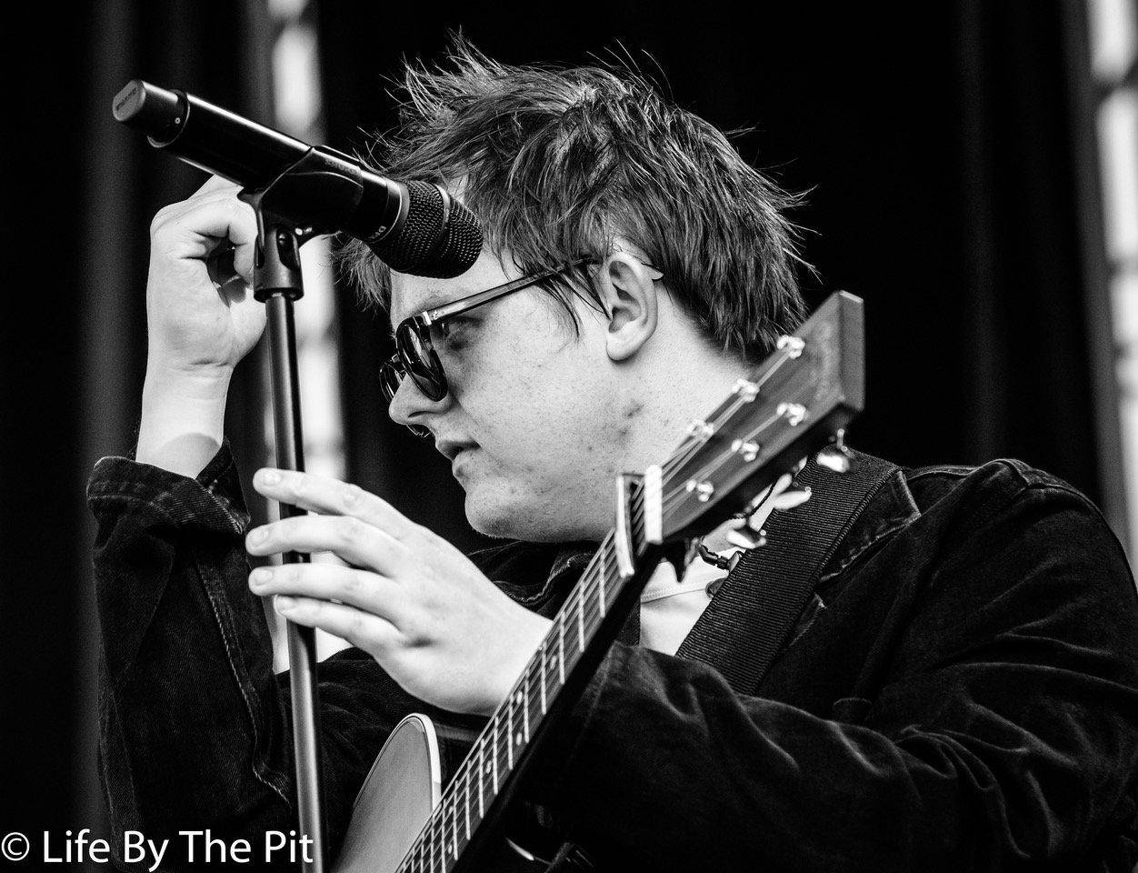 Lewis Capaldi 桌布 壁紙 — 一位戴著太陽眼鏡的音樂家在黑白表演照中調整麥克風，同時彈奏木吉他。