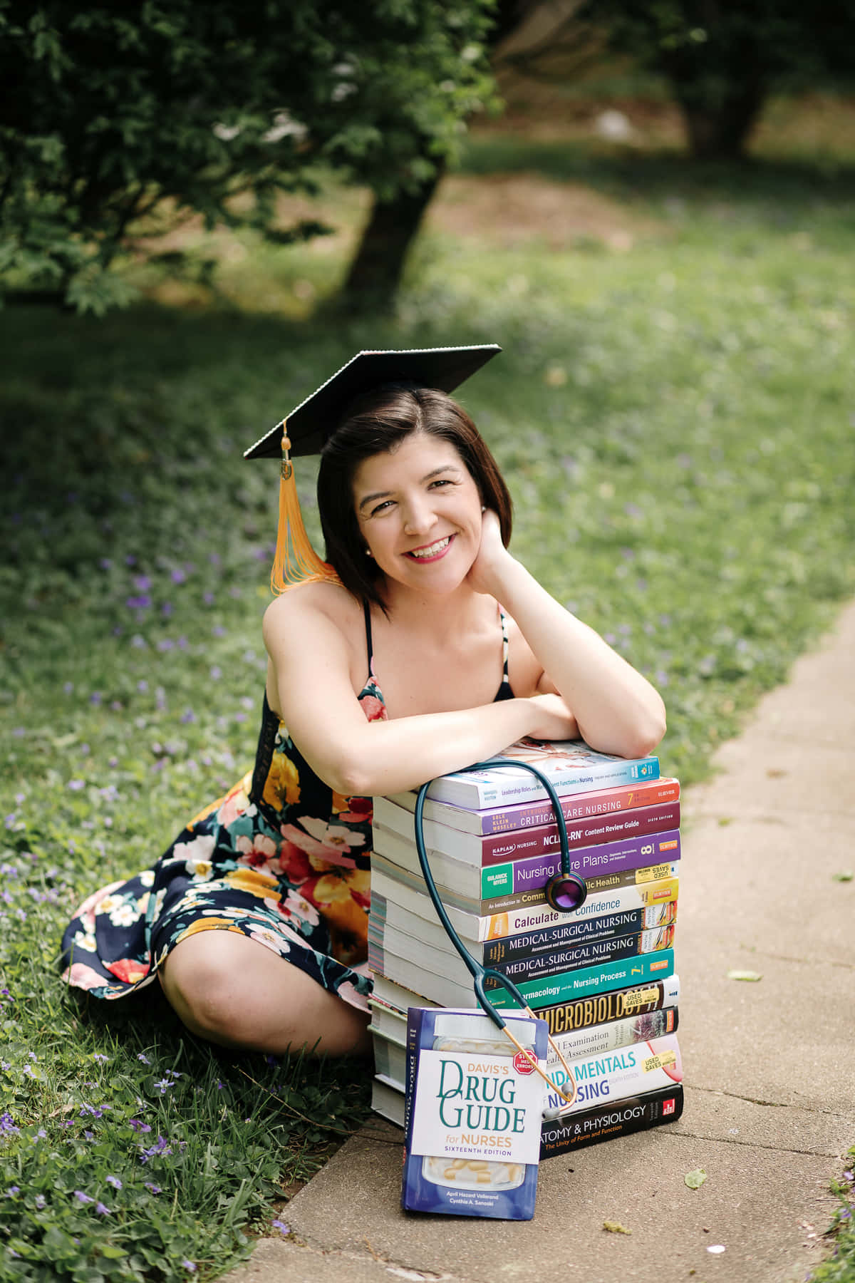 Nurse Graduation 桌布 壁紙 — 一位戴著學士帽、身穿碎花裙的畢業生微笑著，在陽光明媚的草地上，將下巴擱在一疊高聳的護理教科書上。