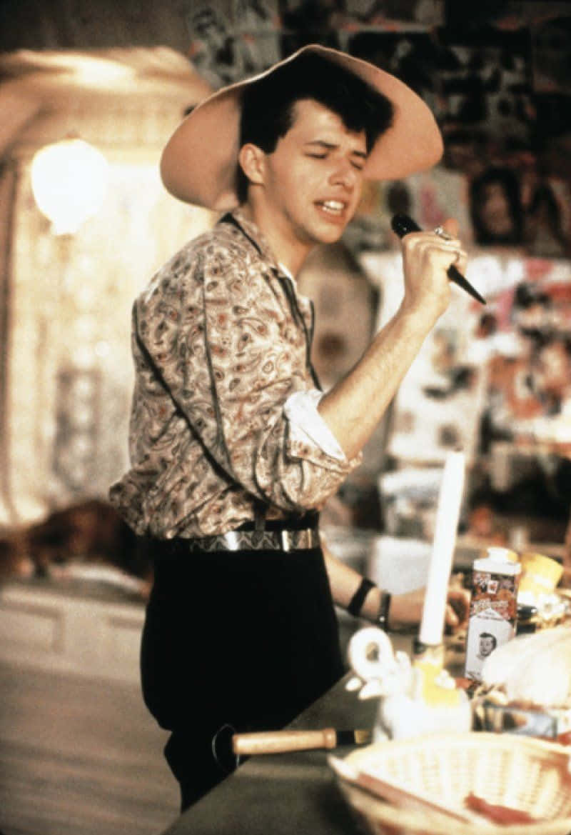 Jon Cryer 桌布 壁紙 — 一位戴著寬邊帽和圖案襯衫的年輕男子在溫暖燈光下、雜亂的房間裡熱情地對著麥克風唱歌。