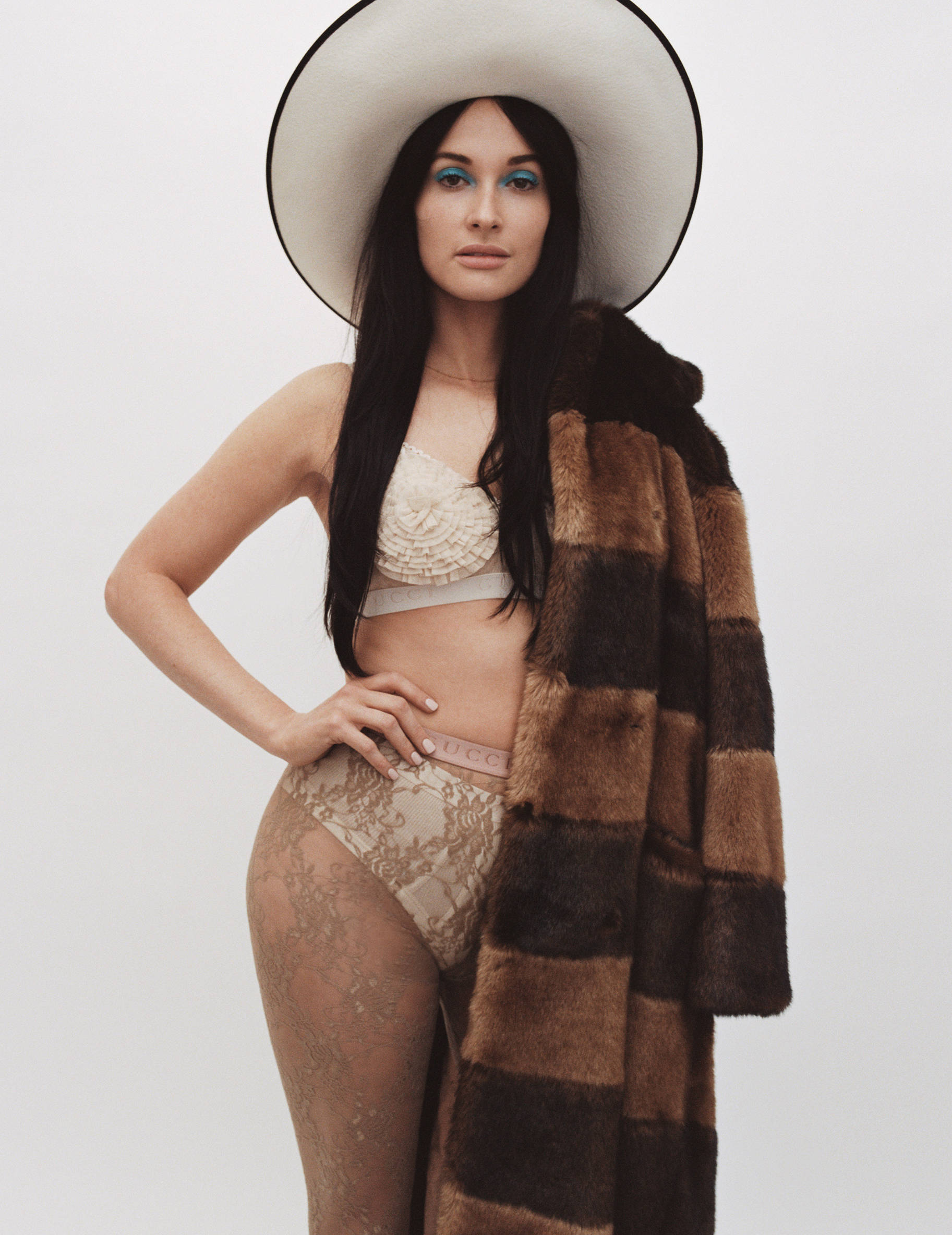 Kacey Musgraves 桌布 壁紙 — 一位戴著寬邊帽和皮草大衣的女性，在純白背景下自信地穿著內衣擺姿勢。