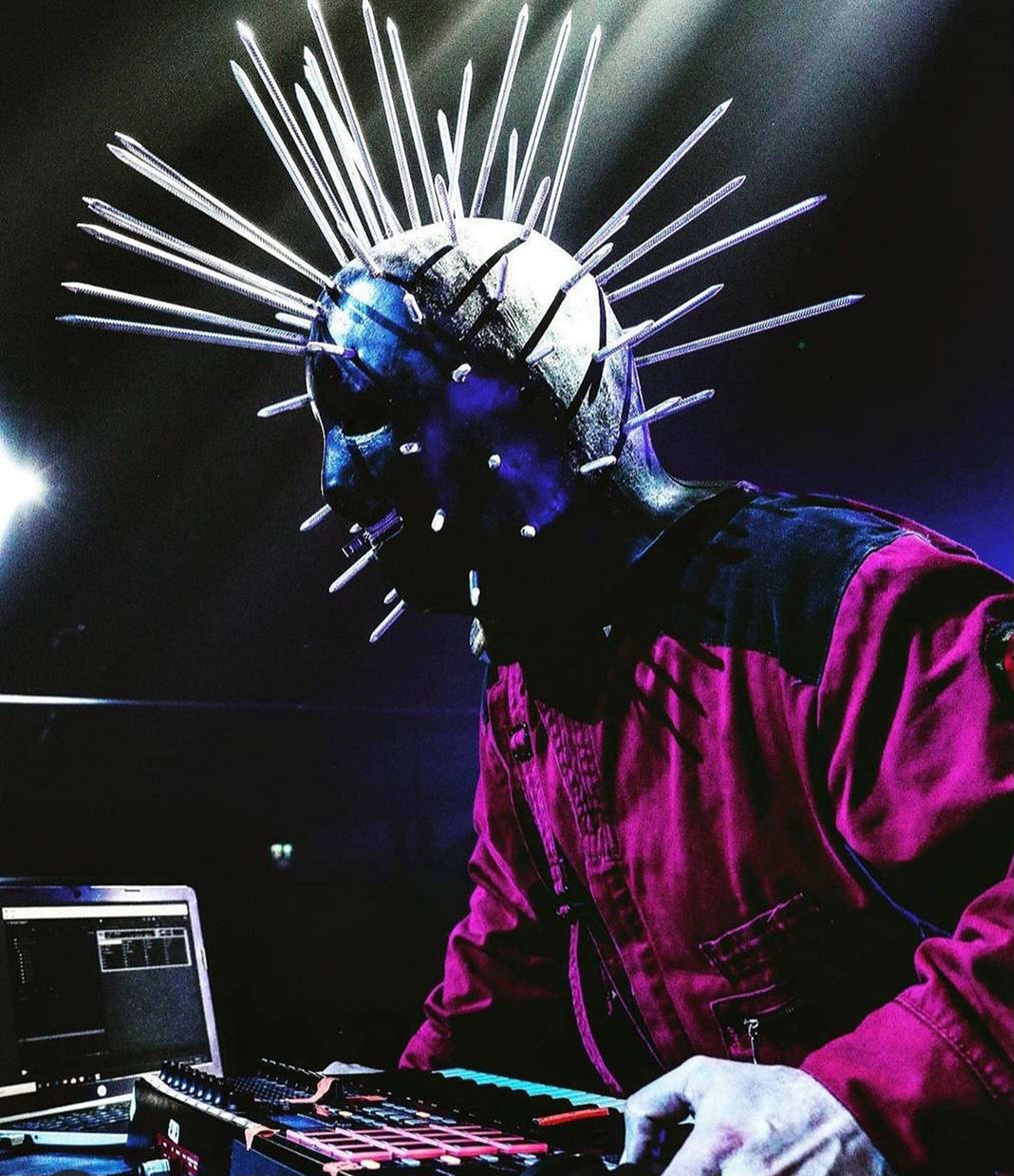 craig jones 桌布 壁紙 — 一位戴著尖刺藍色面具和洋紅色外套的音樂家在黑暗的舞台上，伴隨著發光的設備，進行激烈的表演。