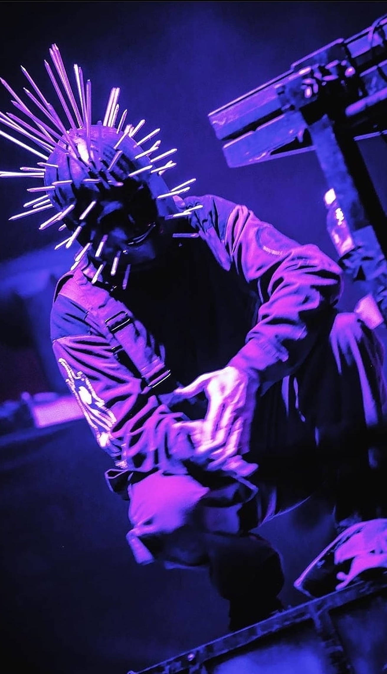 craig jones 桌布 壁紙 — 一位戴著尖刺頭盔的音樂家在黑暗的音樂會場景中，在強烈的紫色舞台燈光下表演。