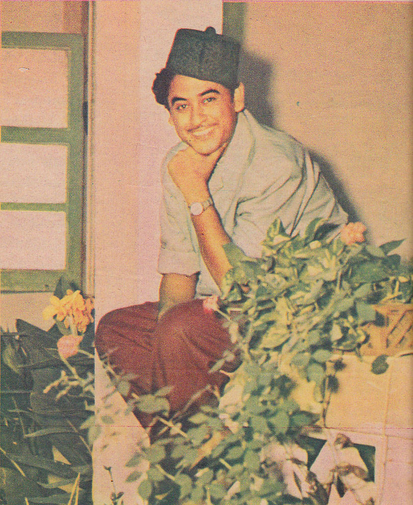Kishore Kumar 桌布 壁紙 — 一位戴著帽子、穿著淺色襯衫的微笑年輕男子，在粉紅色牆壁旁，隨意地坐在茂盛的綠色植物和黃色花朵旁邊。