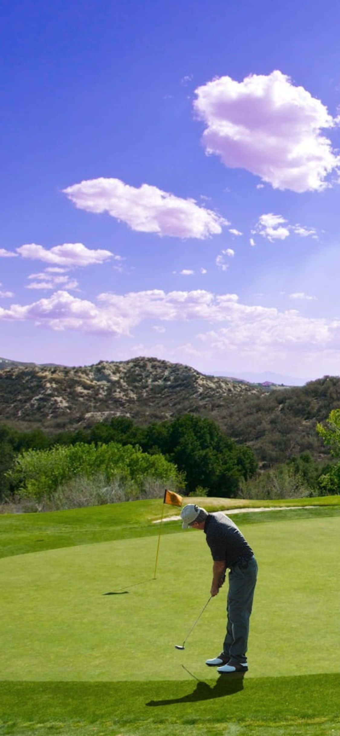 iPhone X Golf Course 桌布 壁紙 — 一位戴著帽子的高爾夫球手在晴朗藍天和蓬鬆白雲下，於充滿活力的果嶺上瞄準推桿。