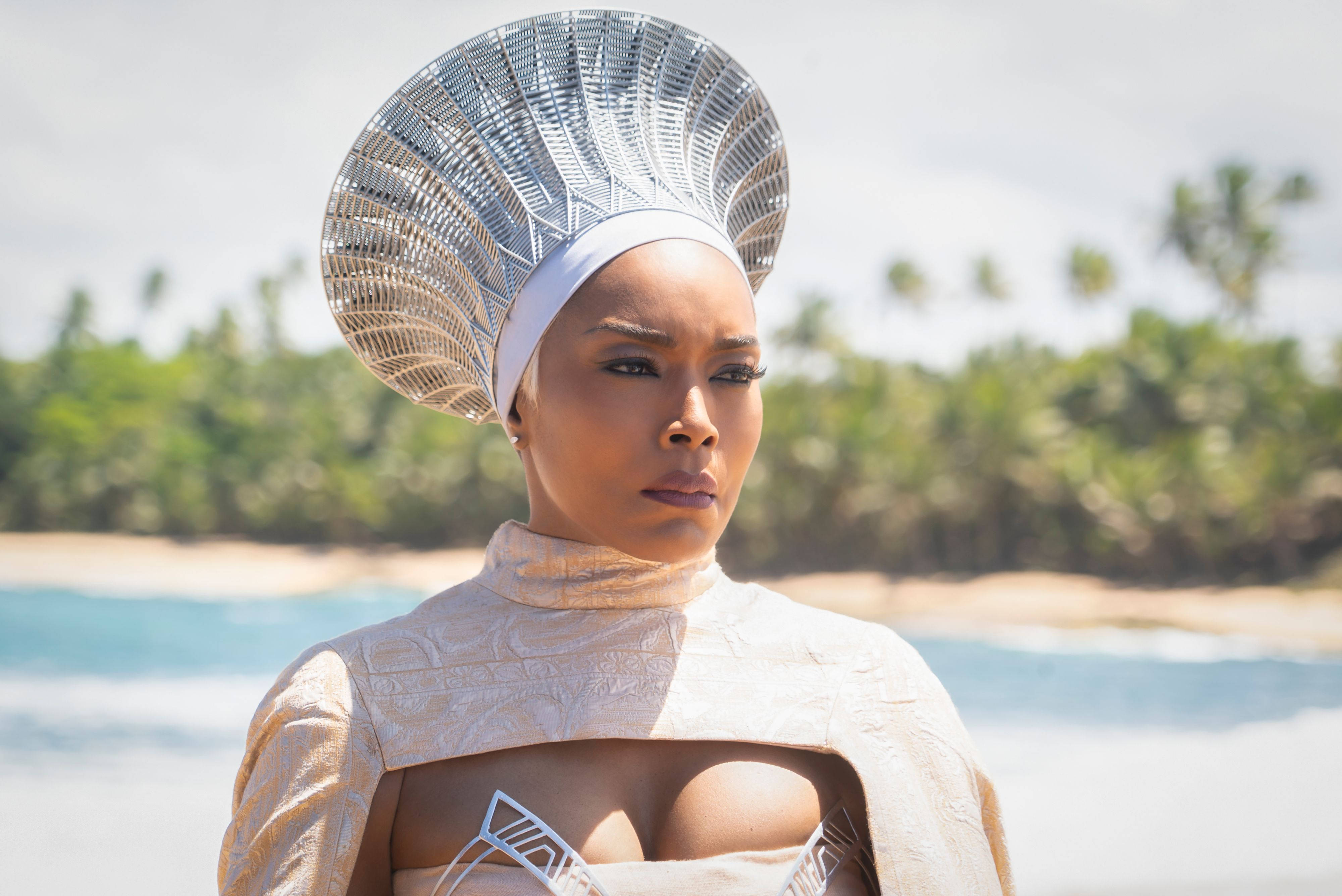 Wakanda 桌布 壁紙 — 一位戴著未來感銀色頭飾、身穿米色服裝的女性，站在陽光明媚的海灘上，身後有棕櫚樹和海洋。