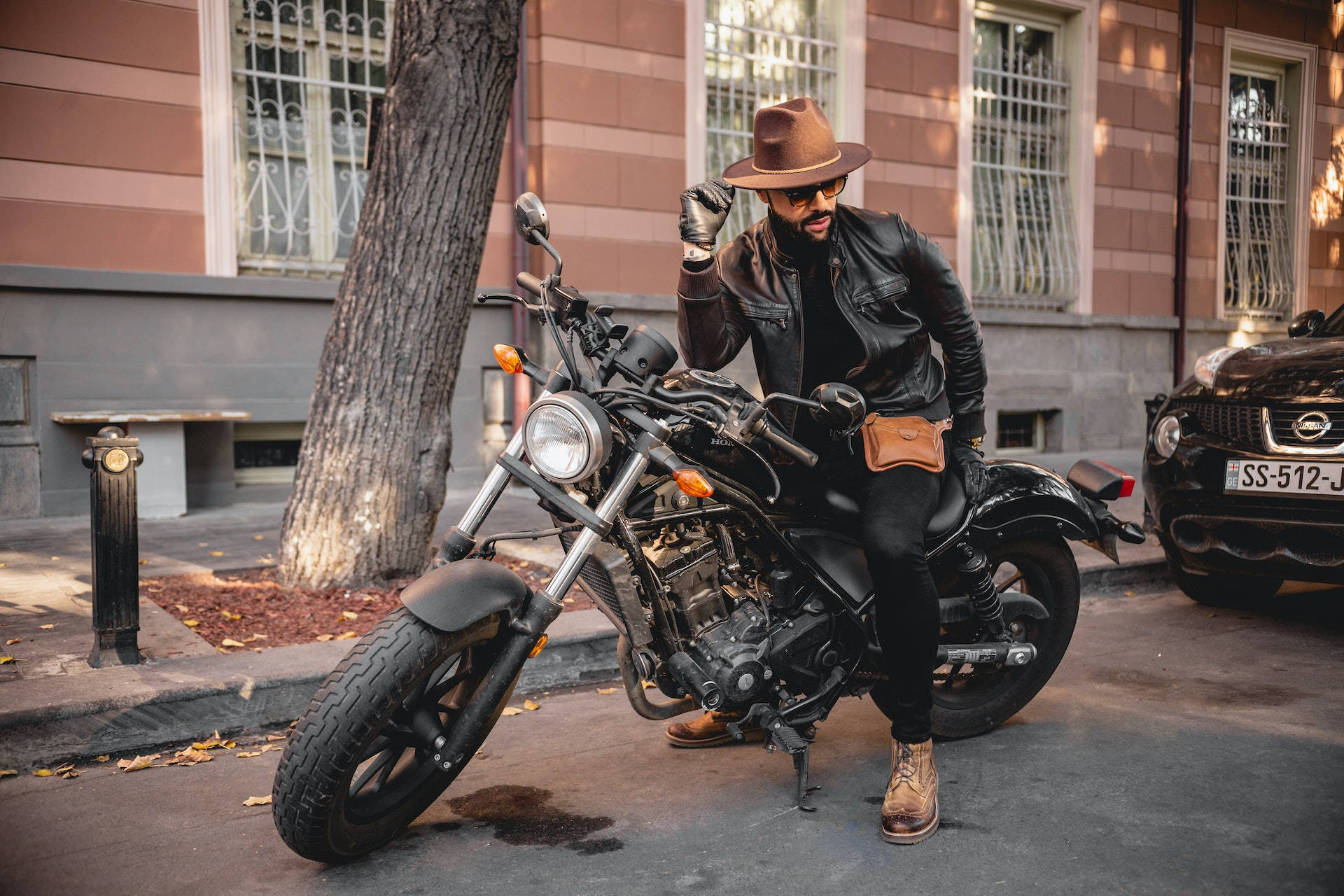 Bike Rider 桌布 壁紙 — 一位戴著棕色帽子和皮夾克的時尚男士，自信地坐在一輛停放在城市人行道上的黑色巡航機車上，旁邊有一棵樹和一座復古建築。