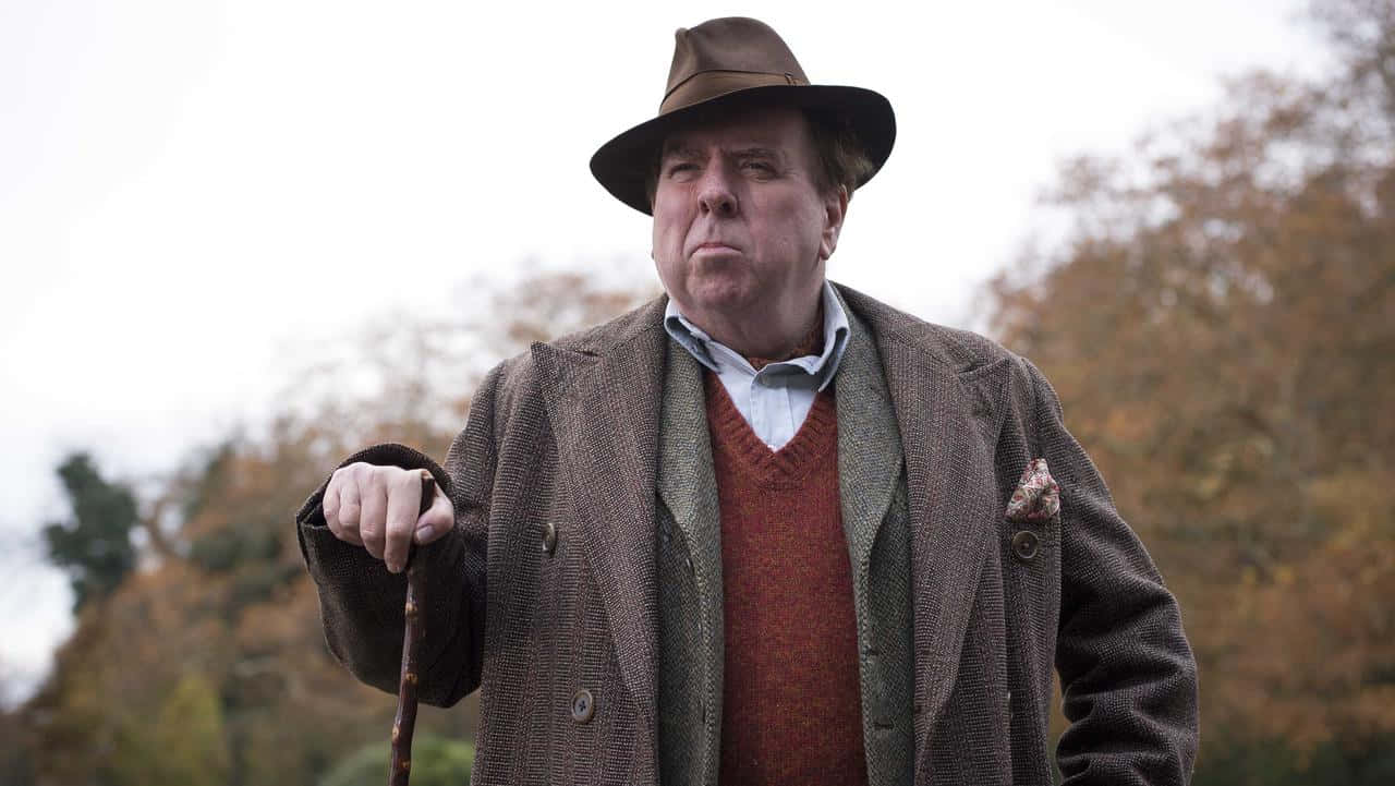 Timothy Spall 桌布 壁紙 — 一位戴著棕色軟呢帽、穿著粗花呢大衣的長者，在金色樹木的秋日背景下手持拐杖。