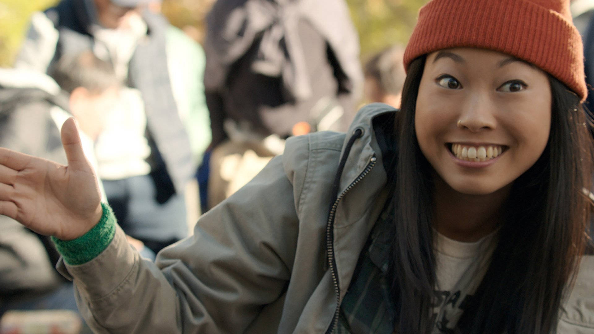 Awkwafina 桌布 壁紙 — 一位戴著橘色毛帽和外套的開朗年輕女性，在陽光明媚的人群中微笑著做手勢。