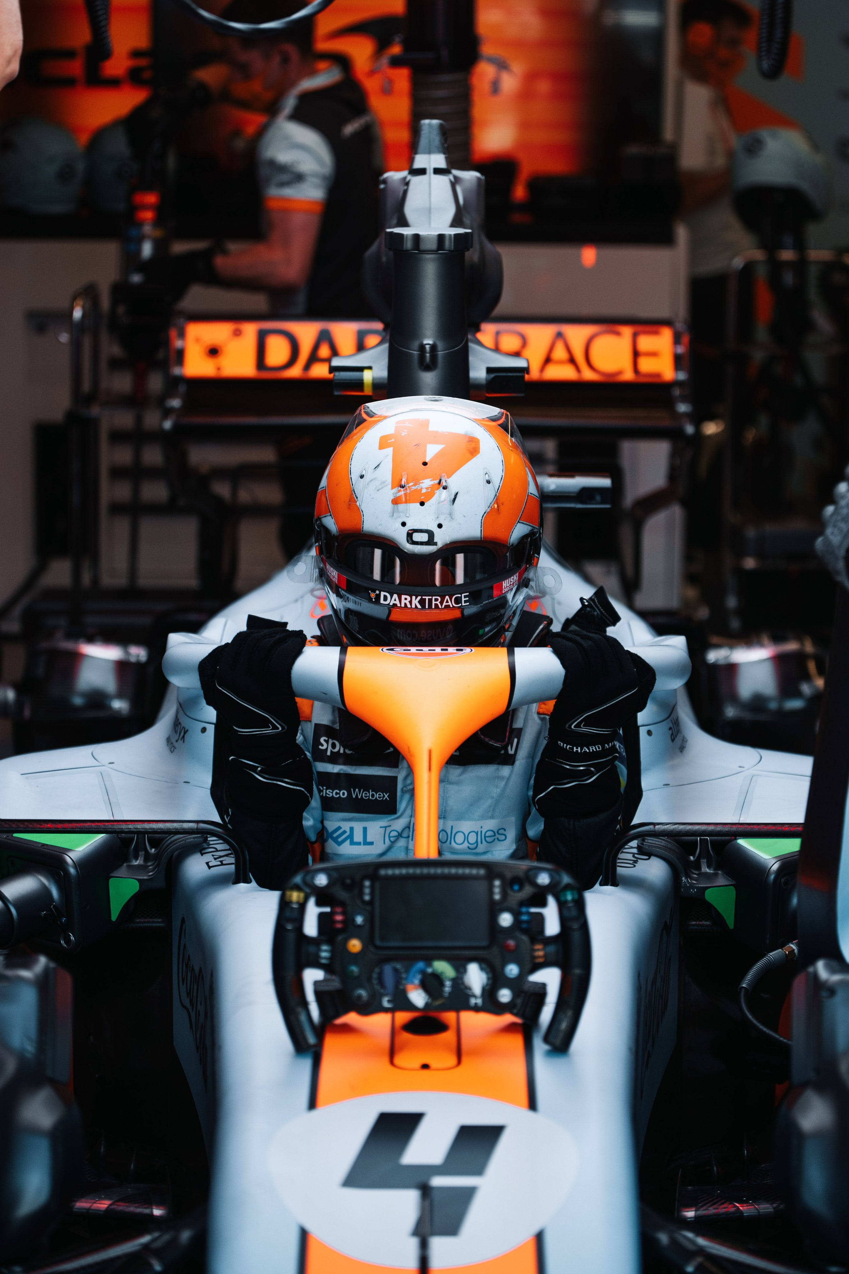 F1 iPhone 壁紙 桌布 壁紙 — 一位戴著橙色和白色頭盔的F1車手，專注地坐在他的賽車內，贊助商標誌清晰可見。