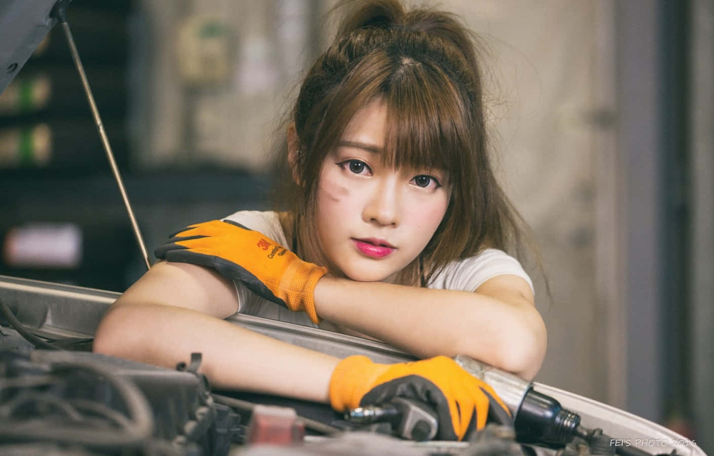 美女 桌布 壁紙 — 一位戴著橙色手套的年輕女子在車庫裡專注地俯身於打開的汽車引擎蓋上。