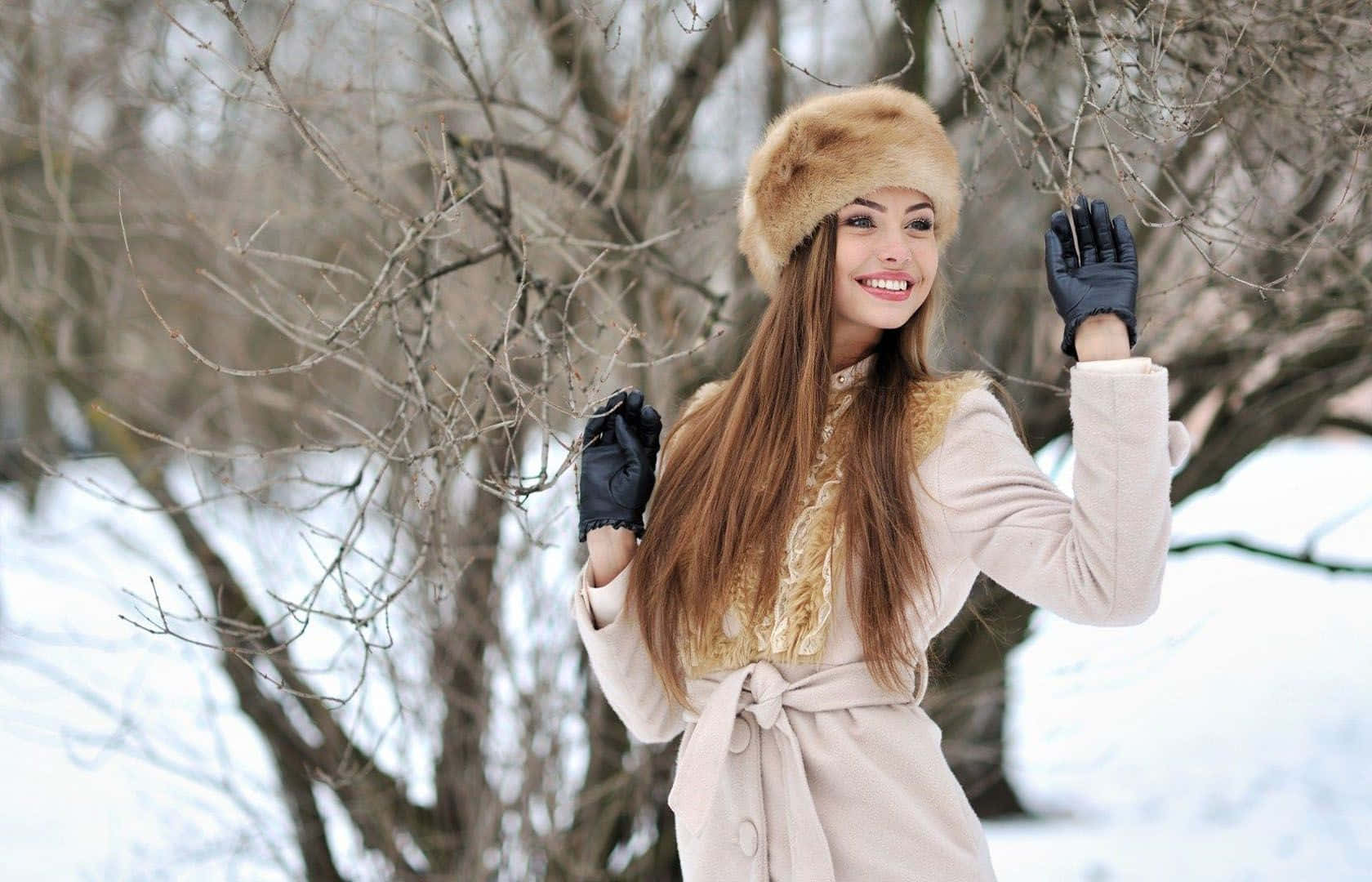 Winter Fashion 桌布 壁紙 — 一位戴著毛帽、身穿米色外套的女士，在雪景背景下頑皮地觸摸光禿禿的冬季樹枝。
