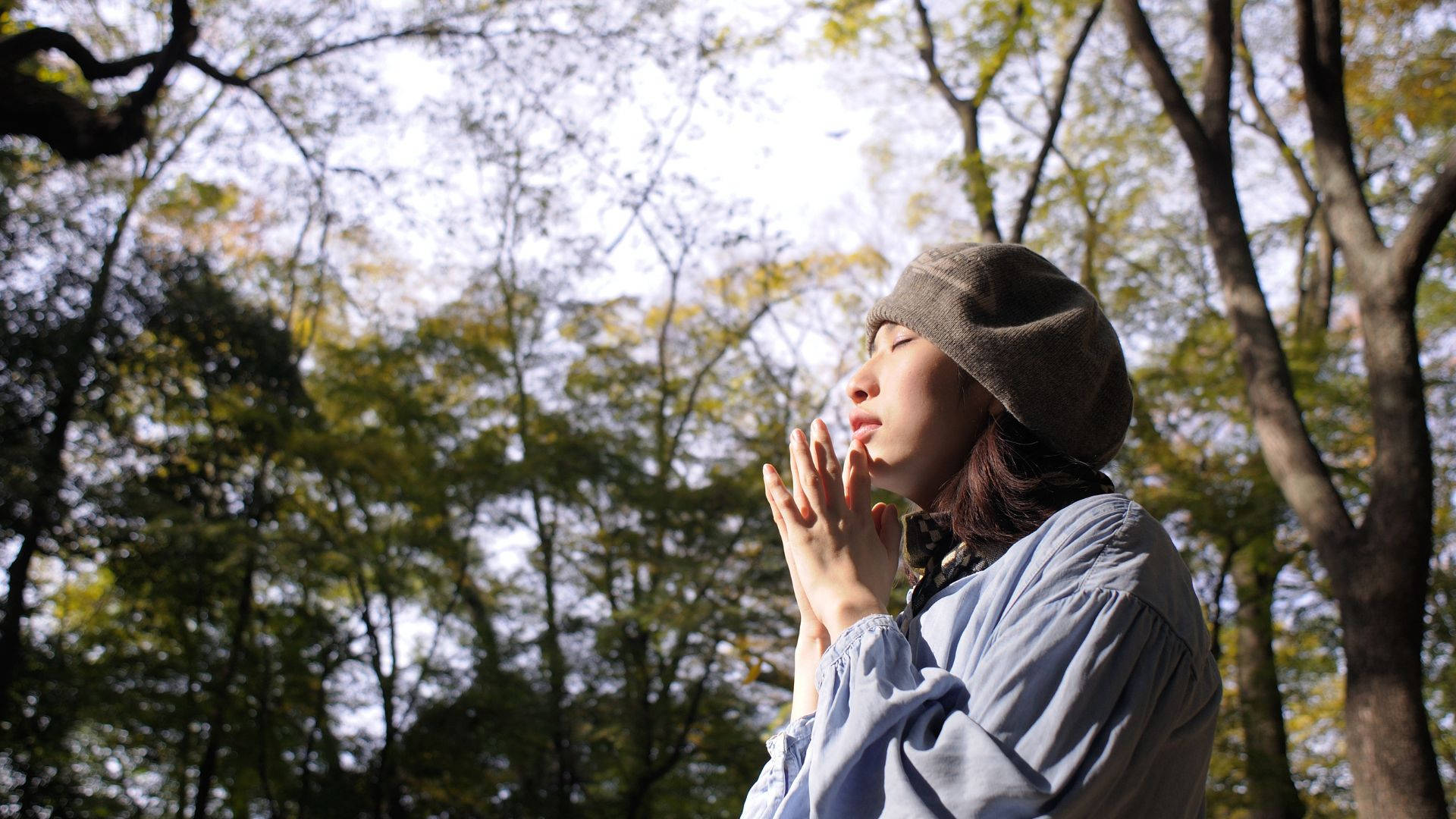 Praying 桌布 壁紙 — 一位戴著毛帽、身穿藍色襯衫的年輕女性，站在陽光普照的秋日森林中，閉上雙眼，雙手合十，靜靜沉思。