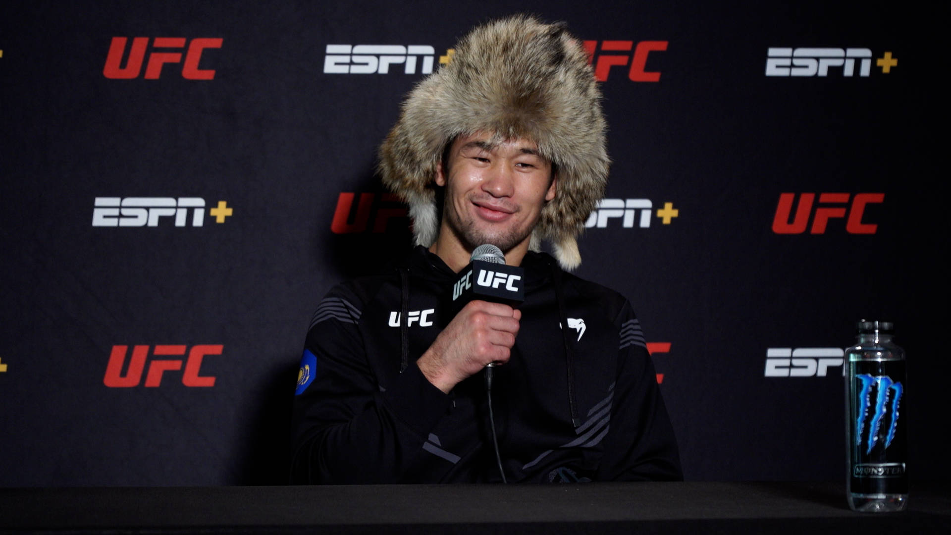 UFC 桌布 壁紙 — 一位戴著毛帽、面帶微笑的拳手在UFC記者會上對著麥克風發言，身後有ESPN+標誌。