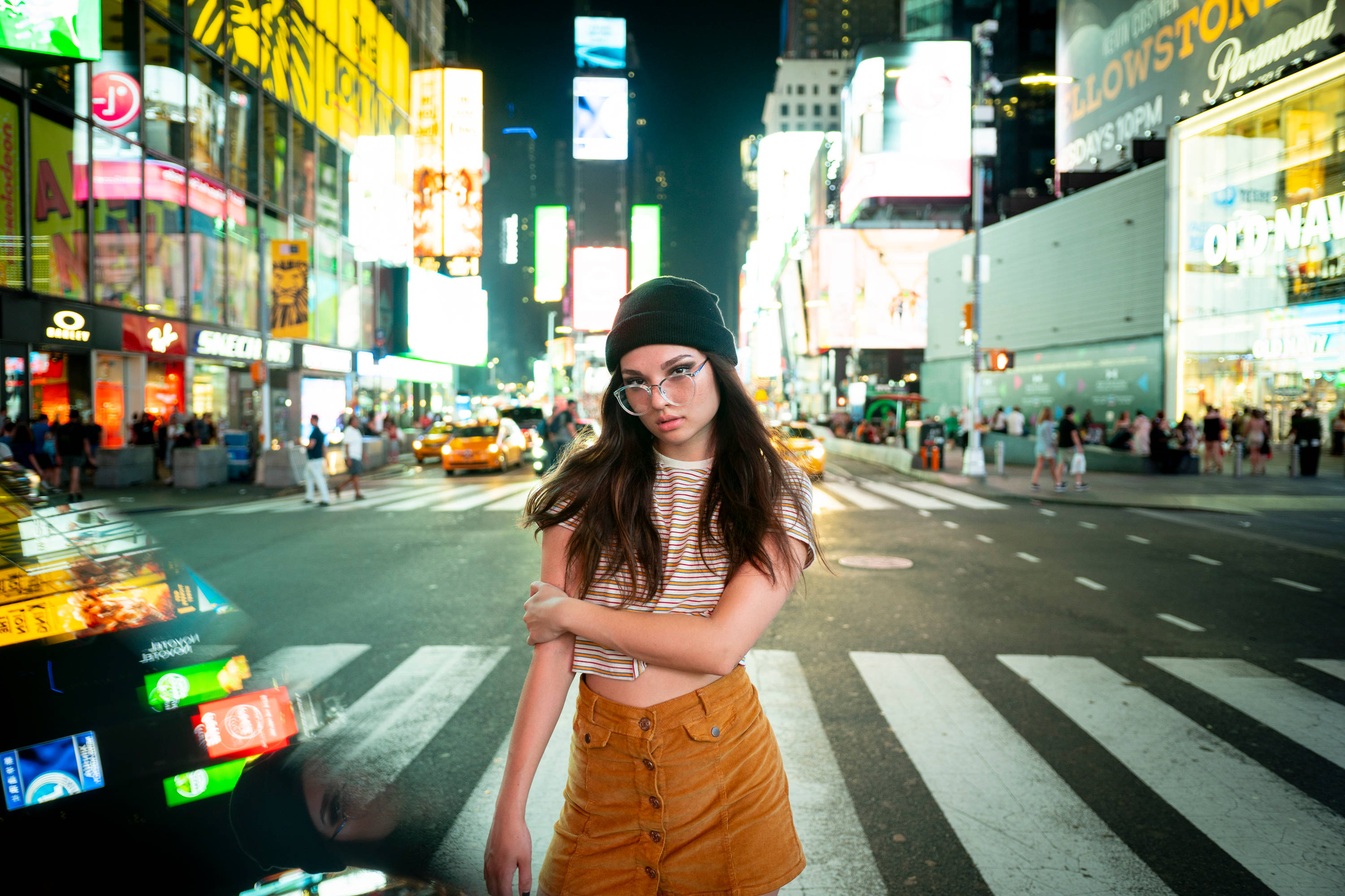 Trendy 桌布 壁紙 — 一位戴著毛帽和眼鏡的年輕女性自信地站在夜晚繁華的霓虹燈街道上，周圍是發光的廣告牌和經過的計程車。