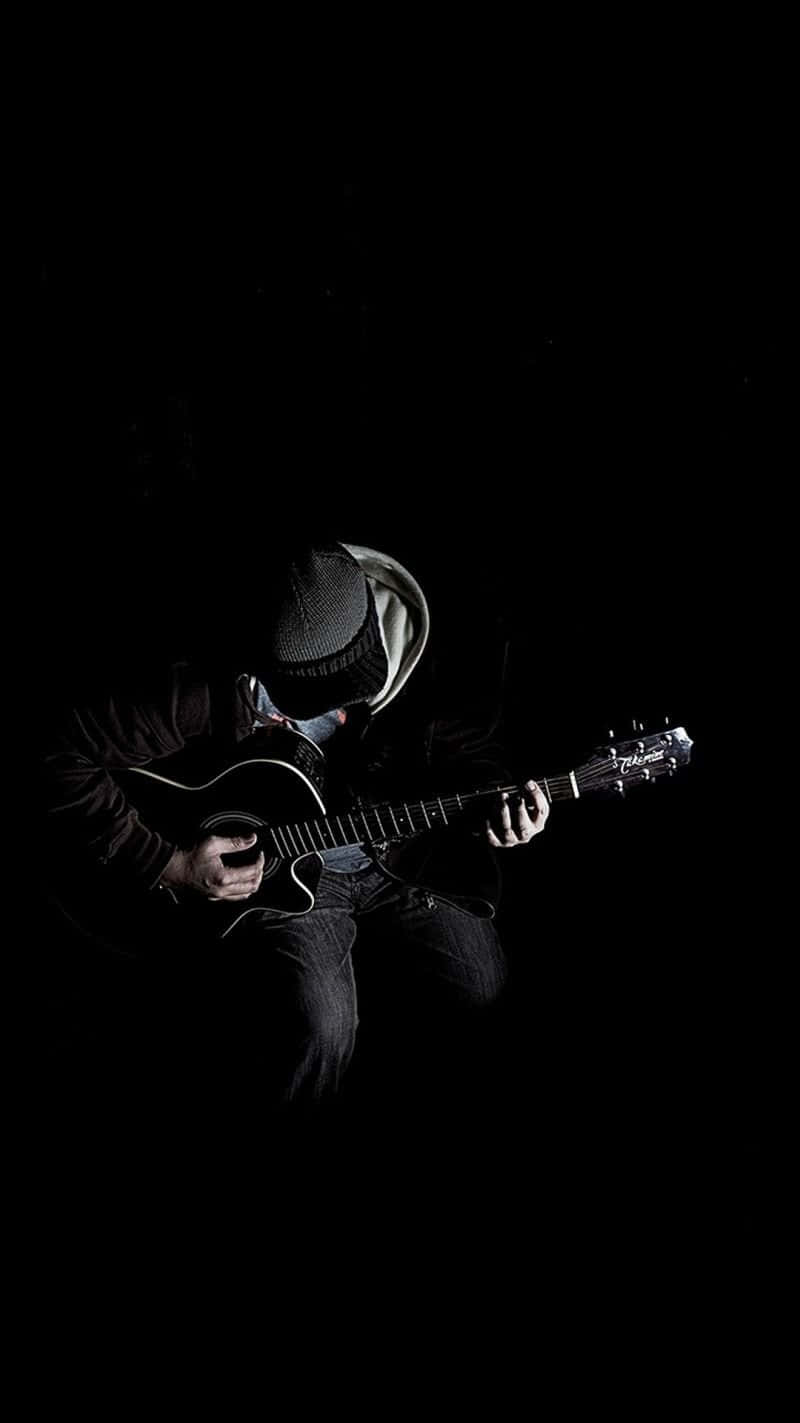 Musical 桌布 壁紙 — 一位戴著毛帽的音樂家在戲劇性的聚光燈下，在漆黑背景前彈奏木吉他。