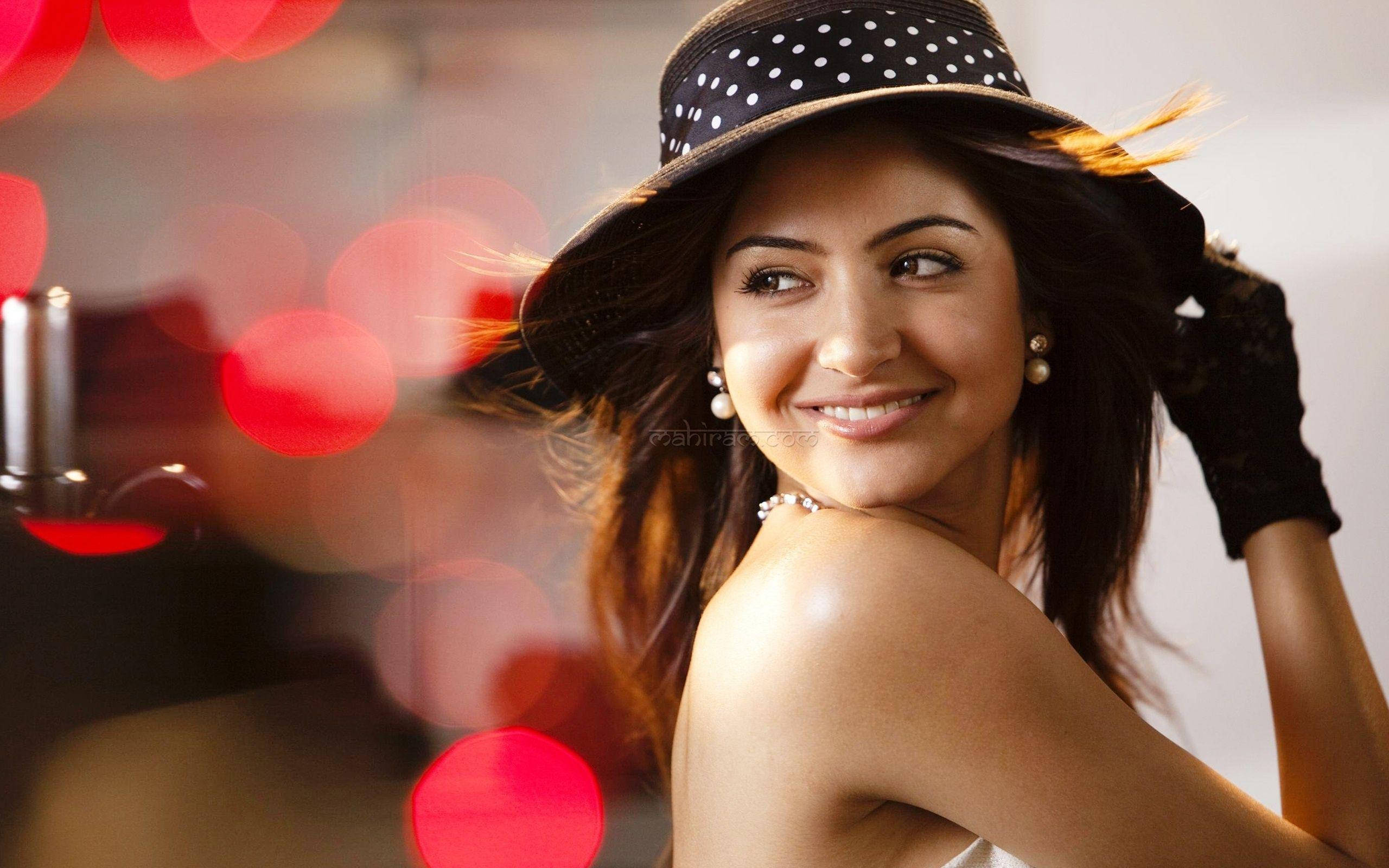 Anushka Sharma 桌布 壁紙 — 一位戴著波點帽和黑色手套的微笑女性，在閃爍的紅色散景燈光背景下擺姿勢。