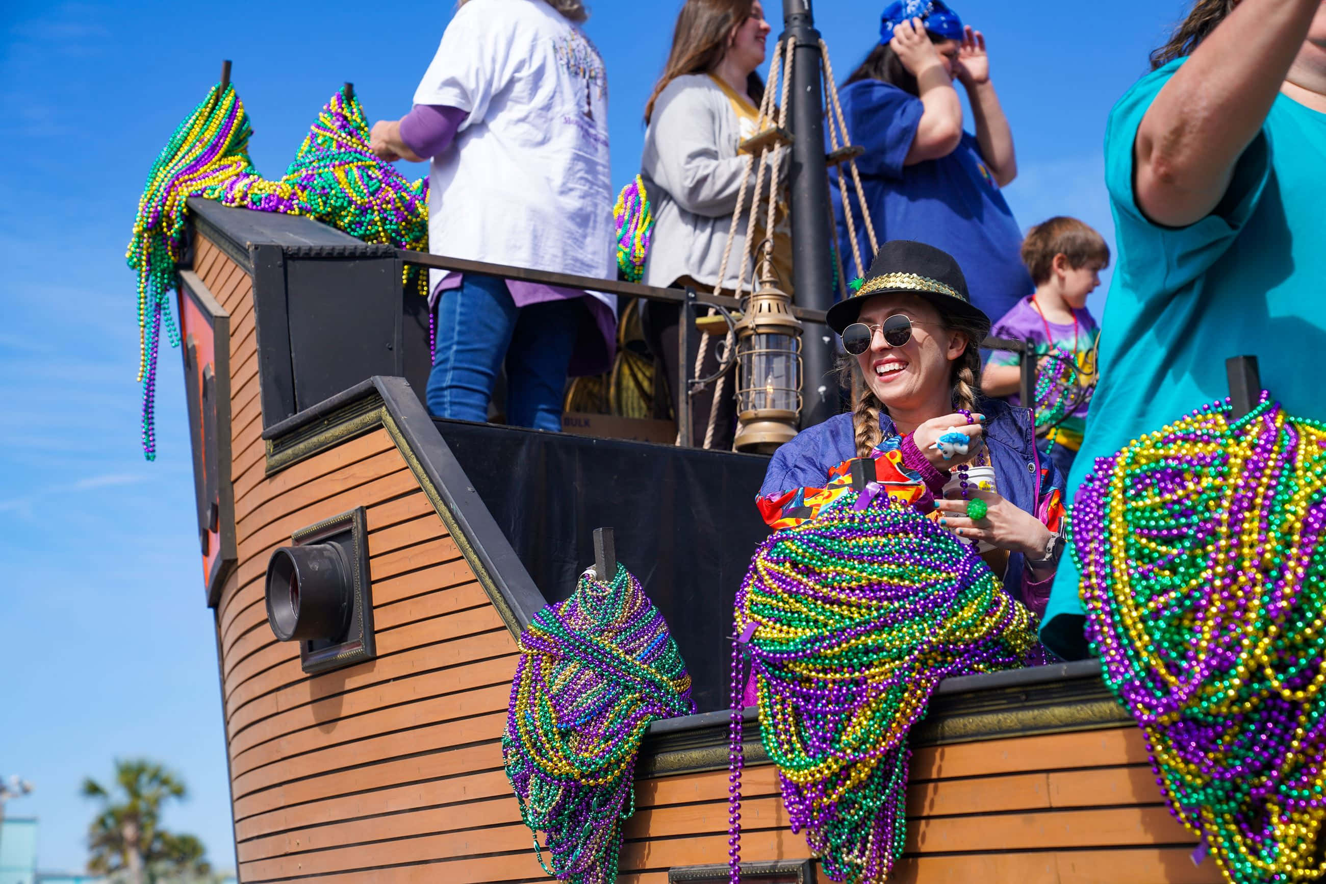 Mardi Gras 桌布 壁紙 — 一位戴著海盜帽和太陽眼鏡的快樂女士，在晴朗的藍天下，在裝飾過的船上拿著色彩繽紛的狂歡節珠子大笑。