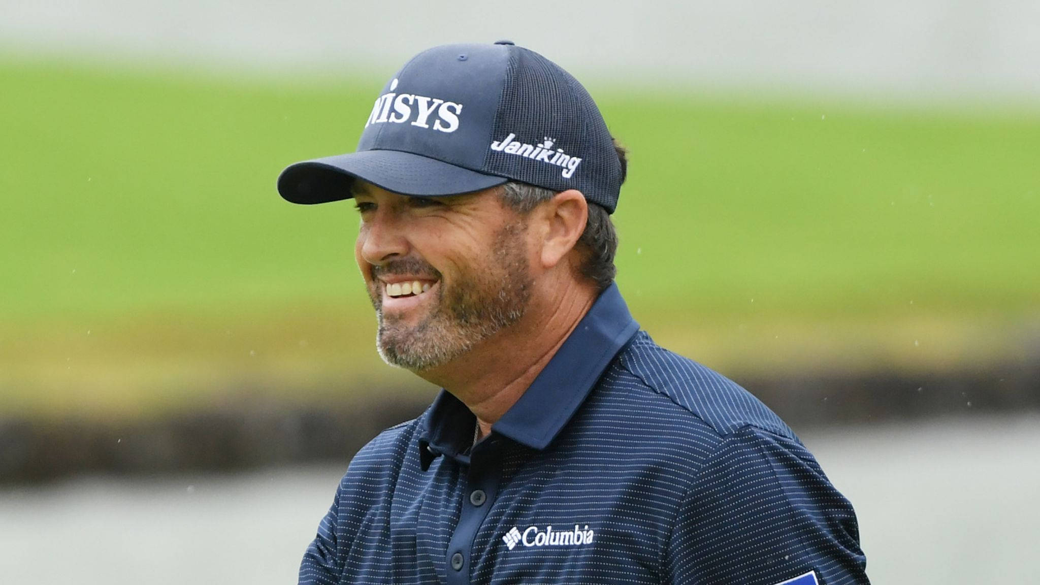 Ryan Palmer 桌布 壁紙 — 一位戴著海軍藍帽子和Polo衫的微笑高爾夫球手站在綠色高爾夫球場上，背景是模糊的草地和水面。