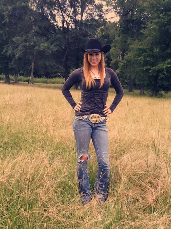 Country Girl 桌布 壁紙 — 一位戴著牛仔帽和破洞牛仔褲的年輕女性自信地站在金色草地上，身後有樹木。