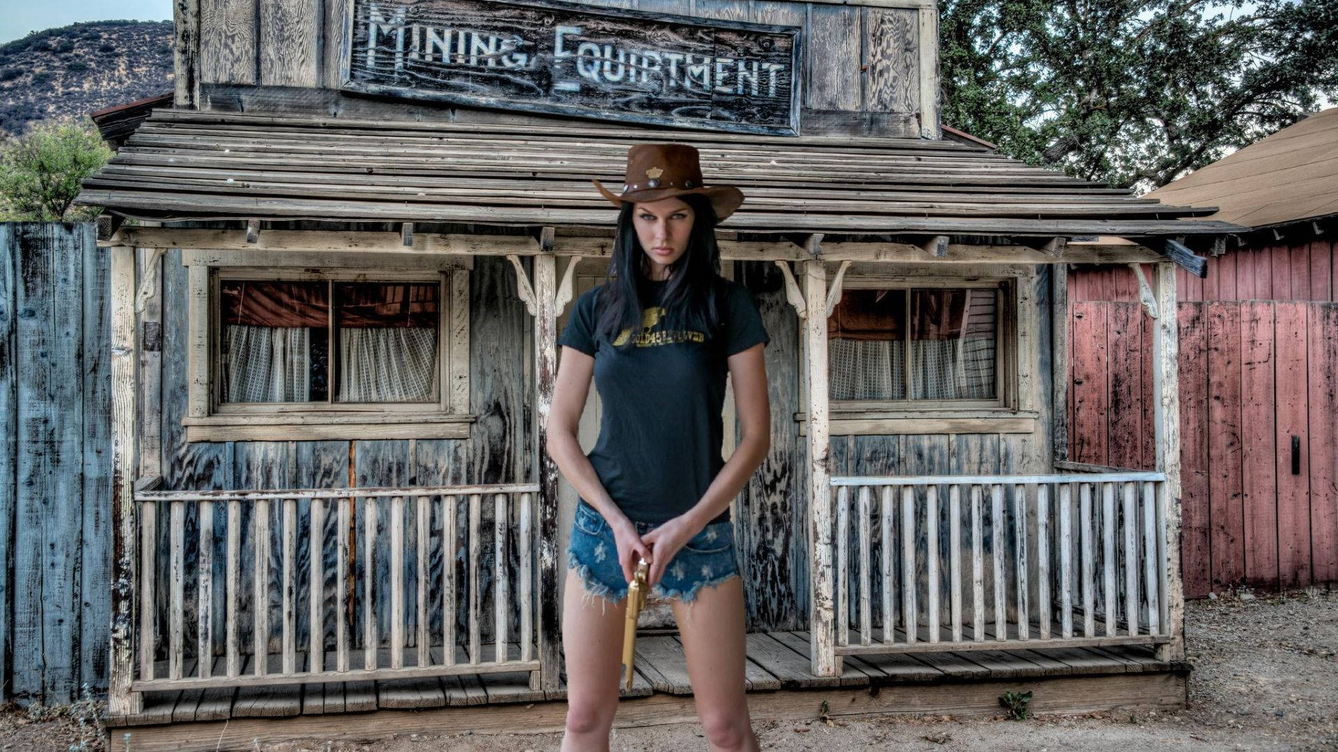 牛仔女孩 桌布 壁紙 — 一位戴著牛仔帽的女性自信地站在一棟寫有「MINING EQUIPMENT」的風化木造建築前，背景是塵土飛揚的西部場景。