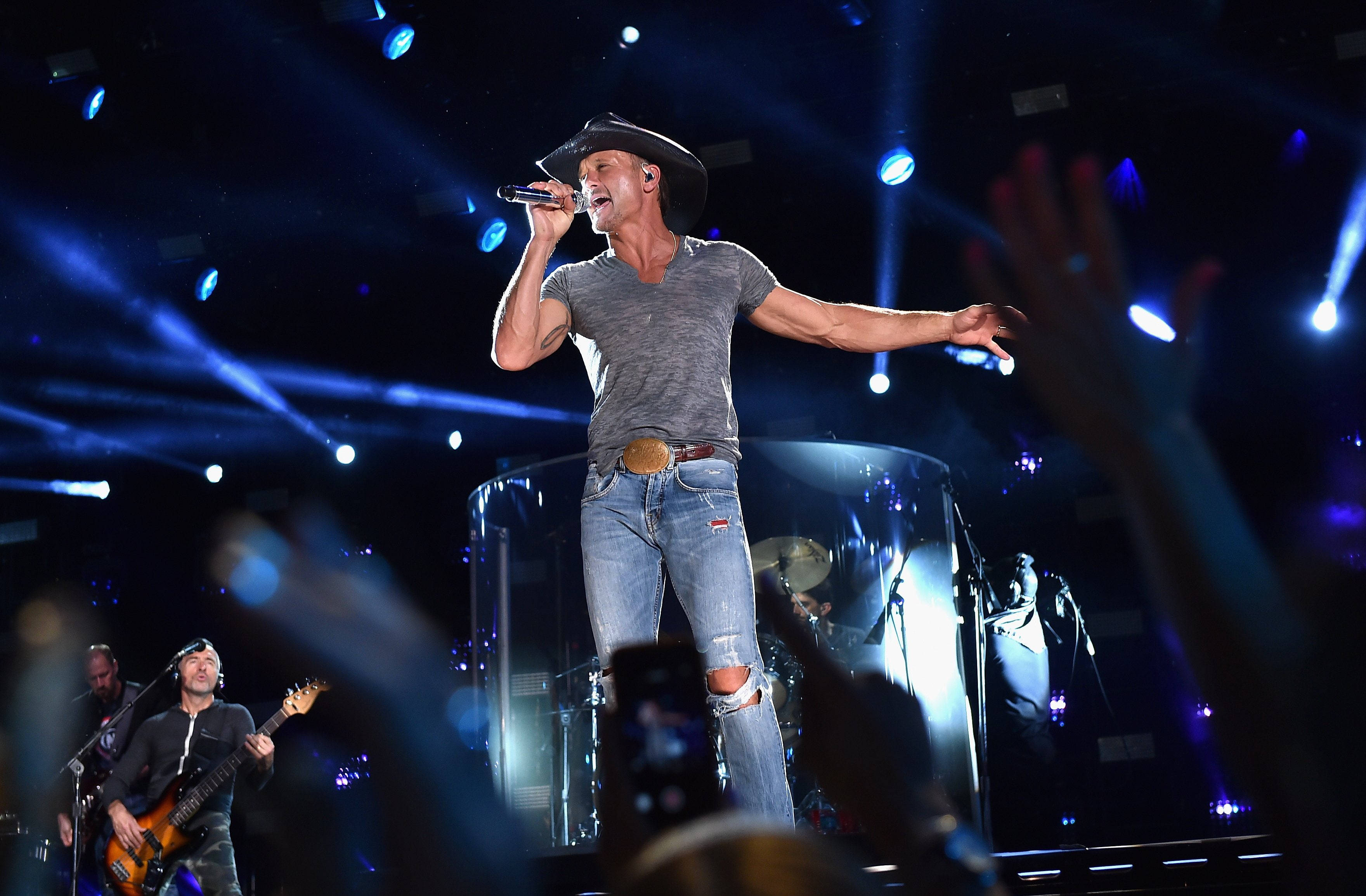 Tim Mcgraw 桌布 壁紙 — 一位戴著牛仔帽的鄉村歌手在藍色燈光的舞台上對著麥克風高歌，粉絲們舉起手和手機。