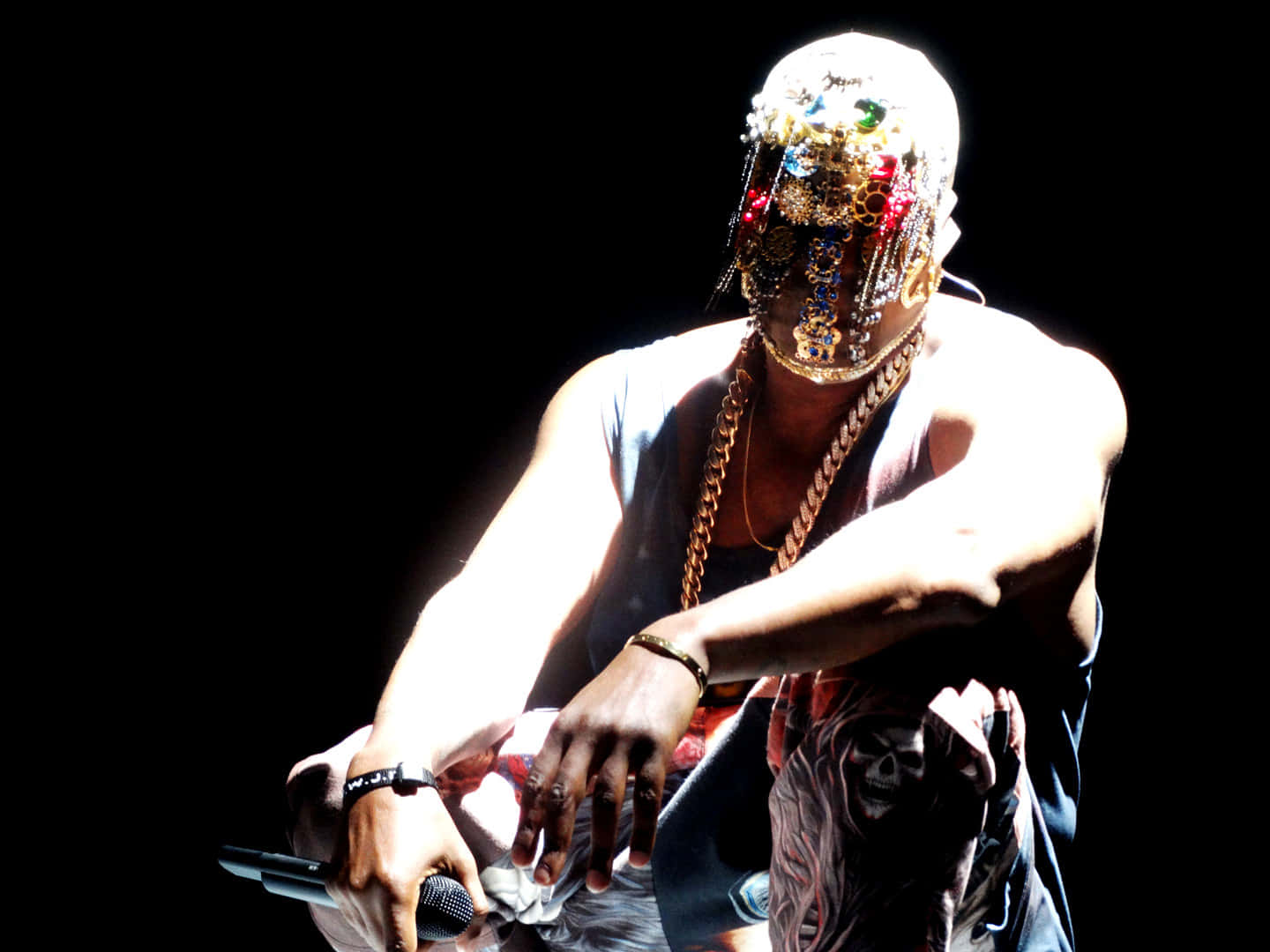 Kanye West Yeezus 桌布 壁紙 — 一位戴著珠寶面具和金鏈的表演者在戲劇性的舞台燈光下拿著麥克風。