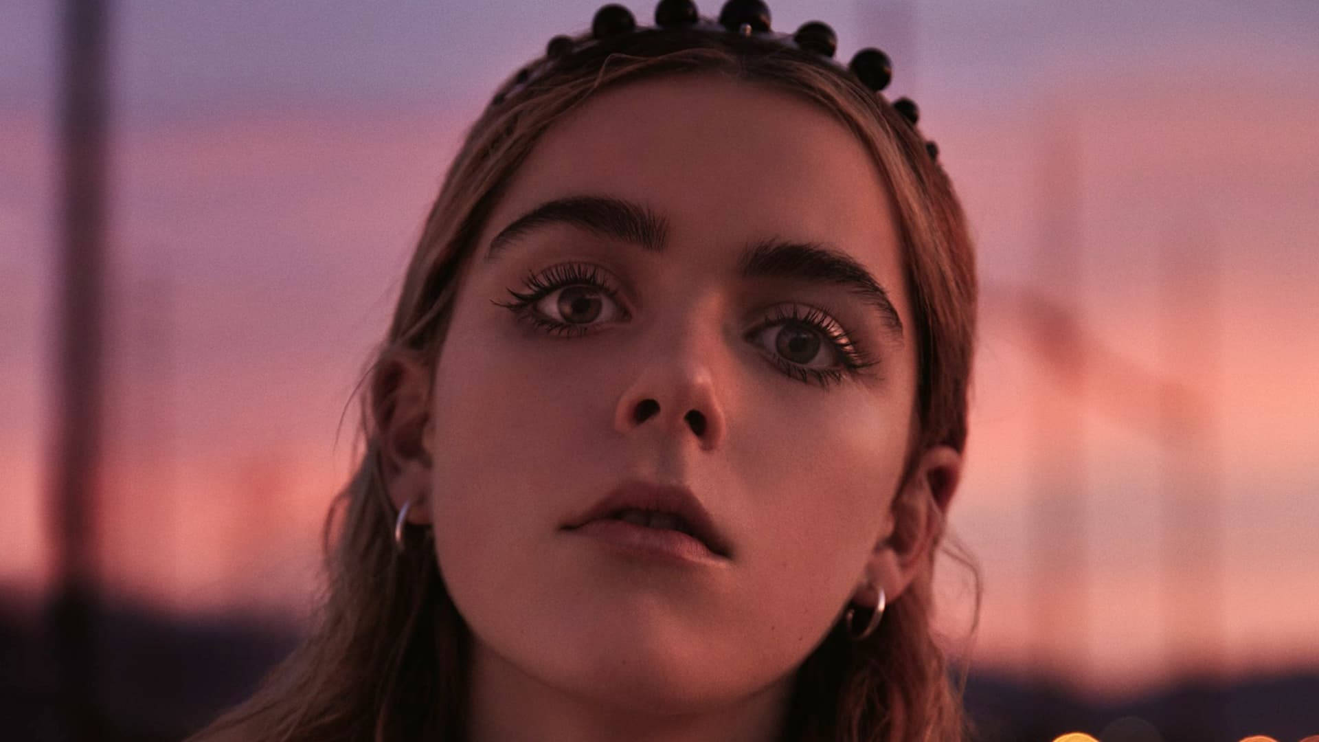 Kiernan Shipka 桌布 壁紙 — 一位戴著珠飾頭帶的年輕女子，在柔和的粉紅與紫色夕陽天空下凝視著。