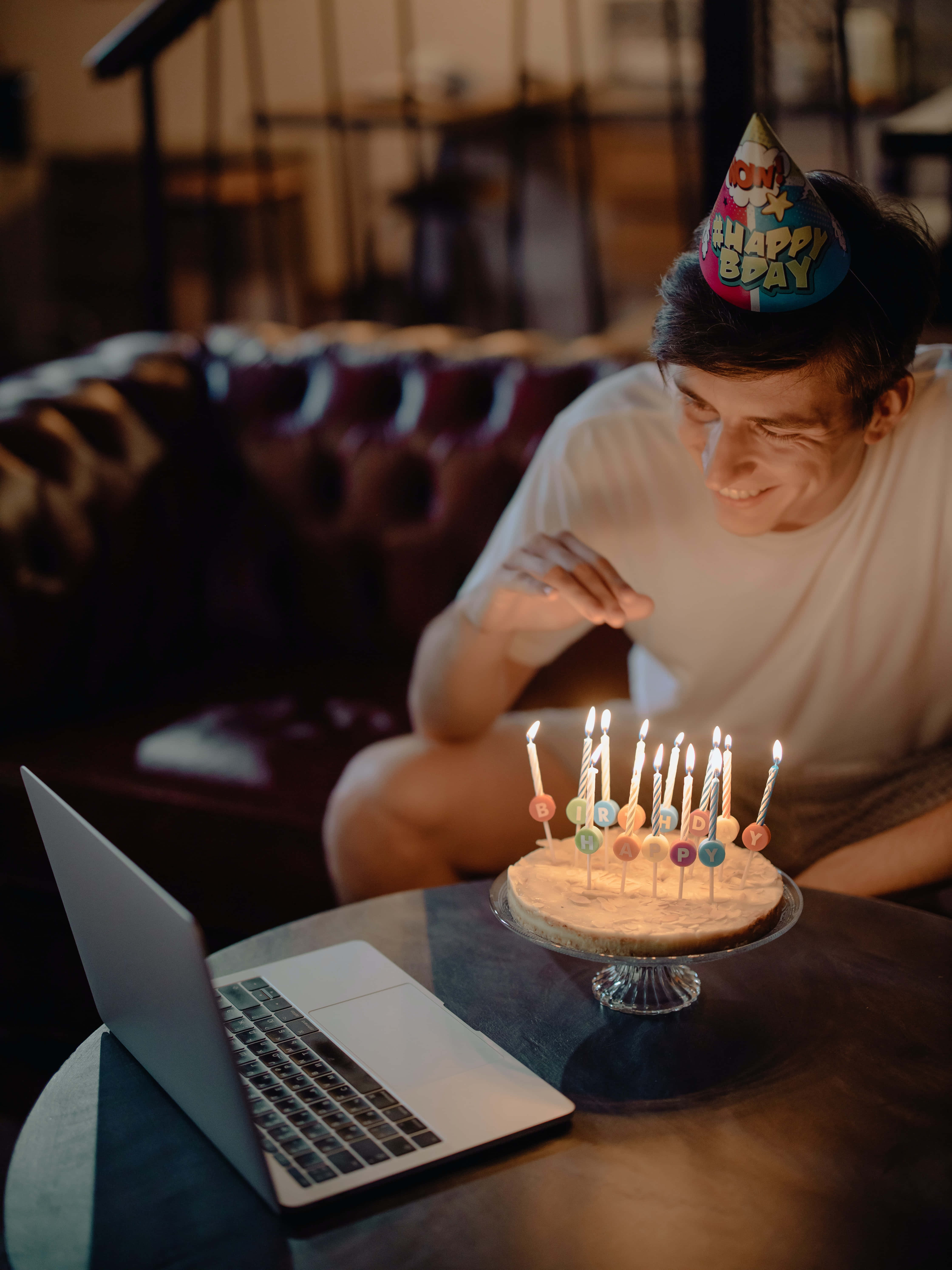 Cute Birthday 桌布 壁紙 — 一位戴著生日帽的年輕男子，一邊在筆記型電腦上進行視訊通話，一邊微笑著吹熄蛋糕上的蠟燭。