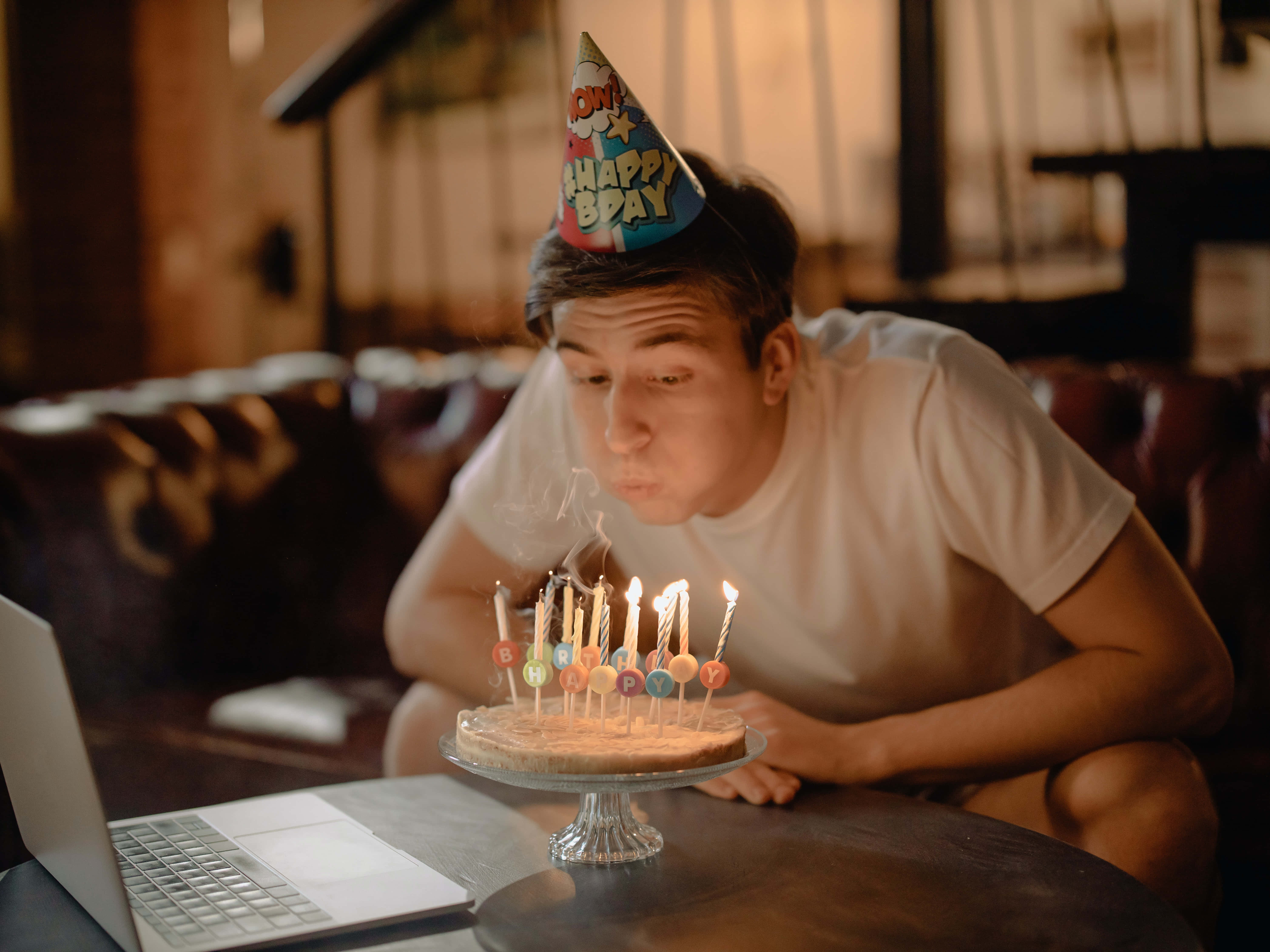生日快樂 桌布 壁紙 — 一位戴著生日帽的年輕男子坐在放有筆記型電腦的桌子前，吹熄蛋糕上的蠟燭。