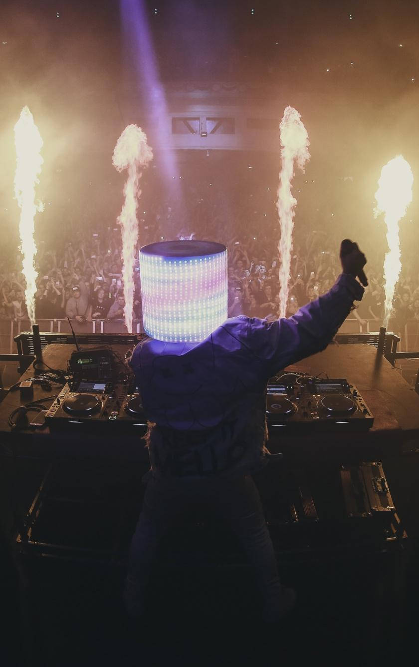 marshmello iPhone 桌布 壁紙 — 一位戴著發光LED頭飾的DJ在舞台上充滿活力地表演，身後有熾熱的煙火爆炸。